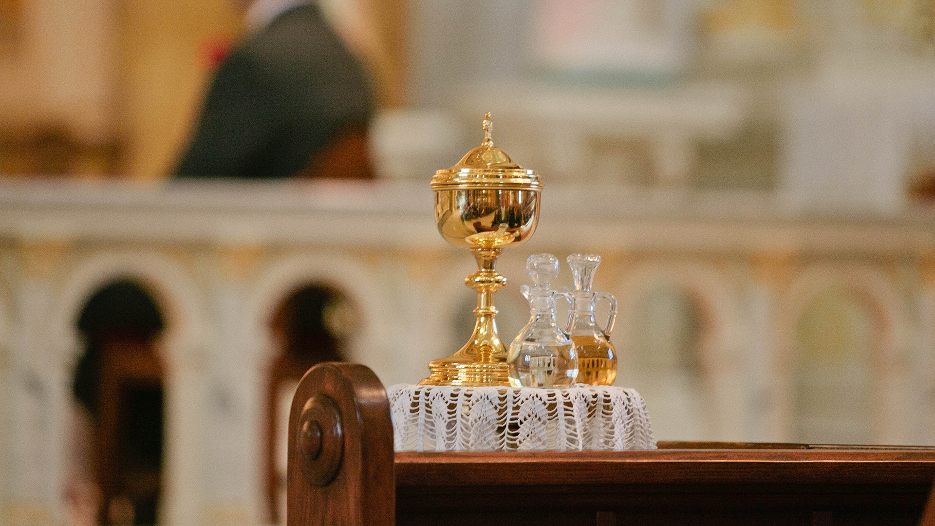 A brass ciborium and two glass cruets on a lace-covered wooden table in a church setting.