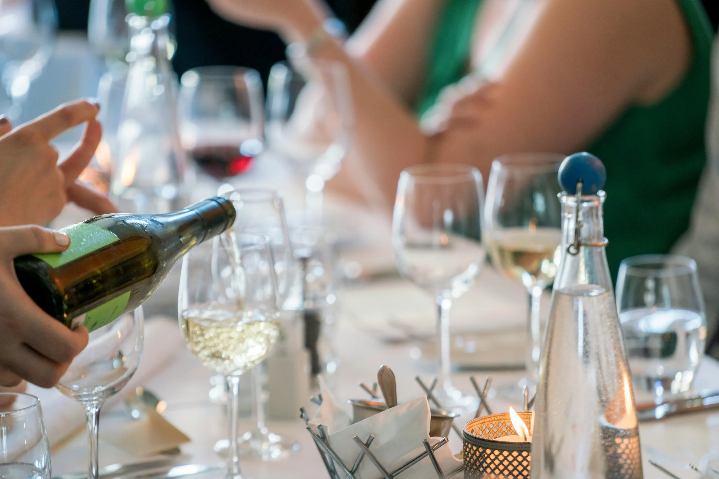 Person pouring white wine into a glass at a table set for a meal, with multiple wine glasses and a lit candle in the foreground.