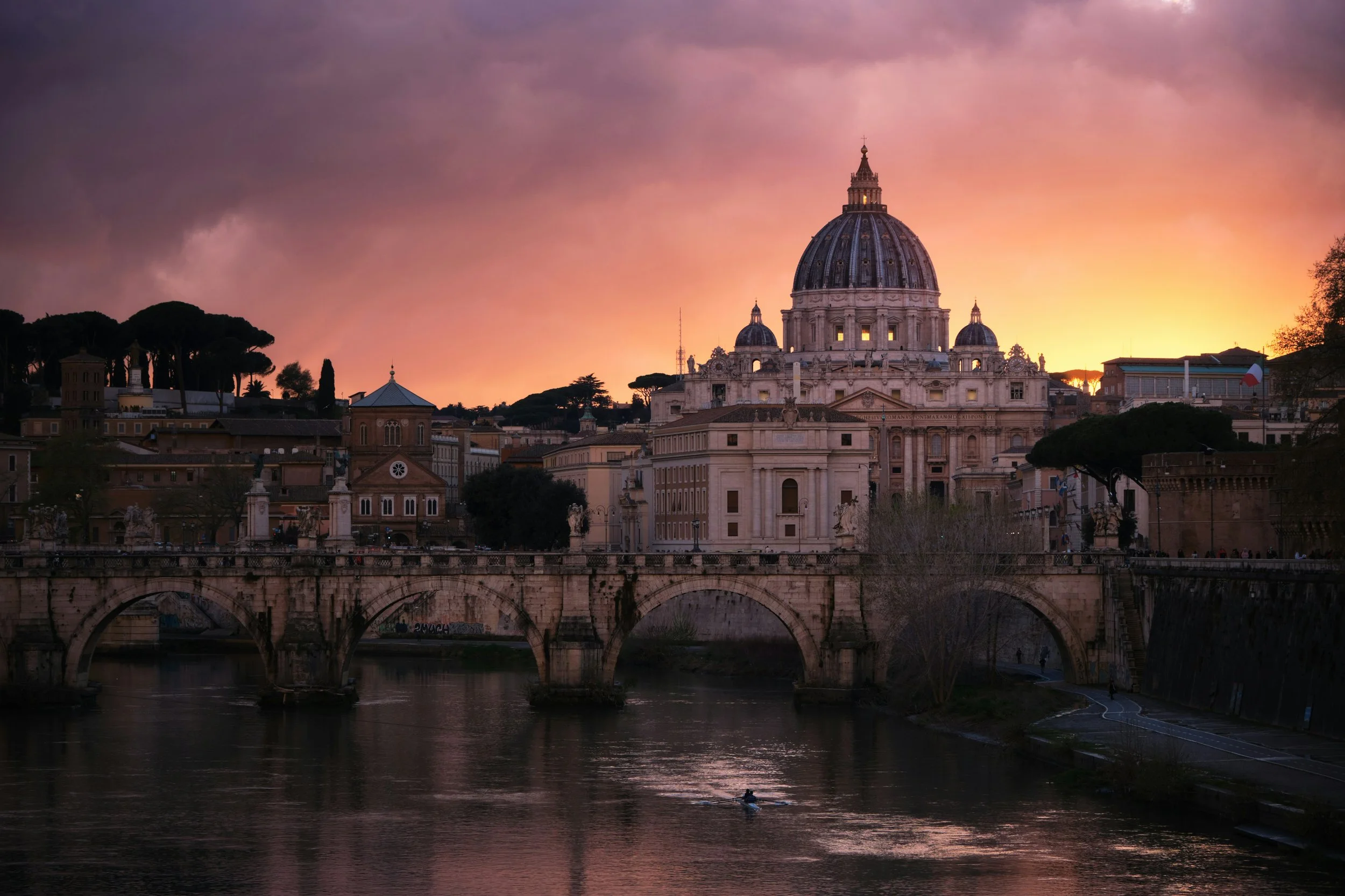 View of St. Peter's Basilica in Vatican City during sunset, with dark clouds in the sky and a river with a person kayaking in the foreground.