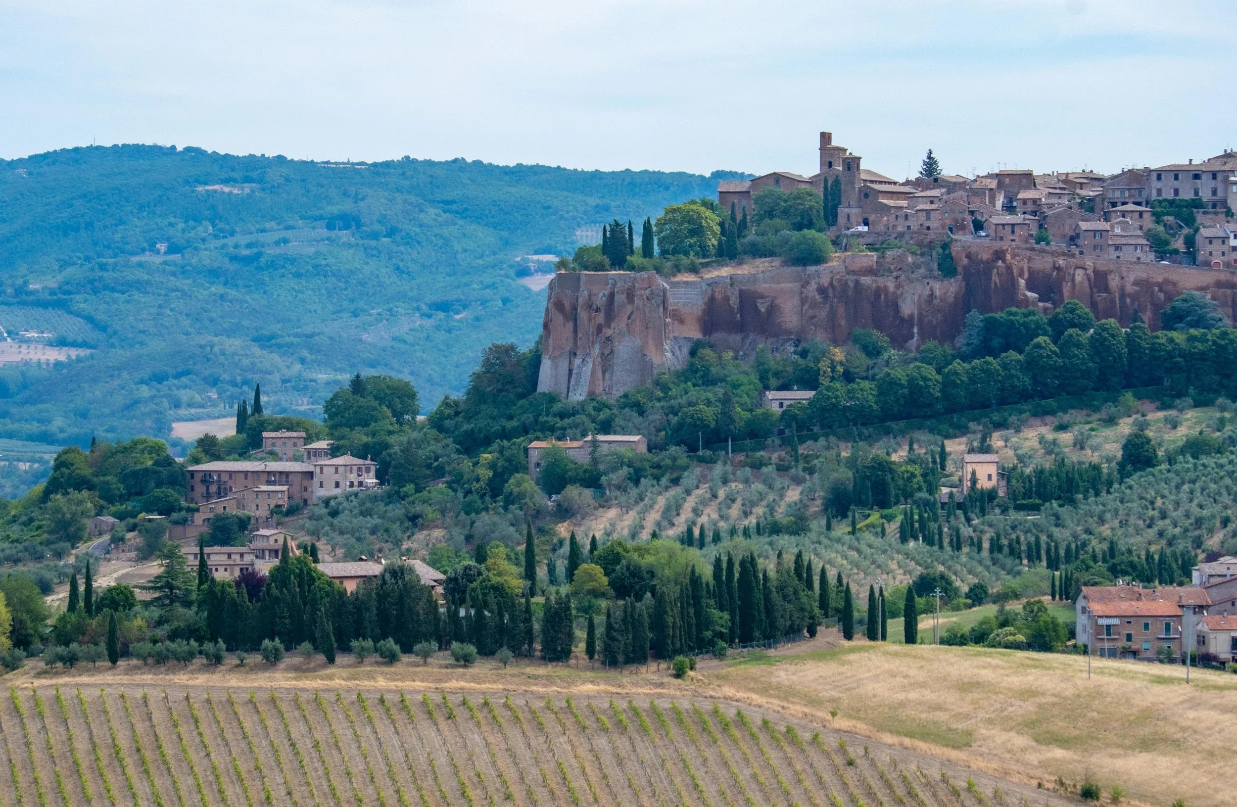 A picturesque hillside village with stone buildings and a church, surrounded by lush green trees and vineyards, set against rolling hills in the background.