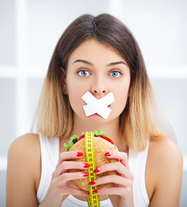 Woman holding a cheeseburger with tape over her mouth symbolizing extreme dieting and starving yourself during fat loss.