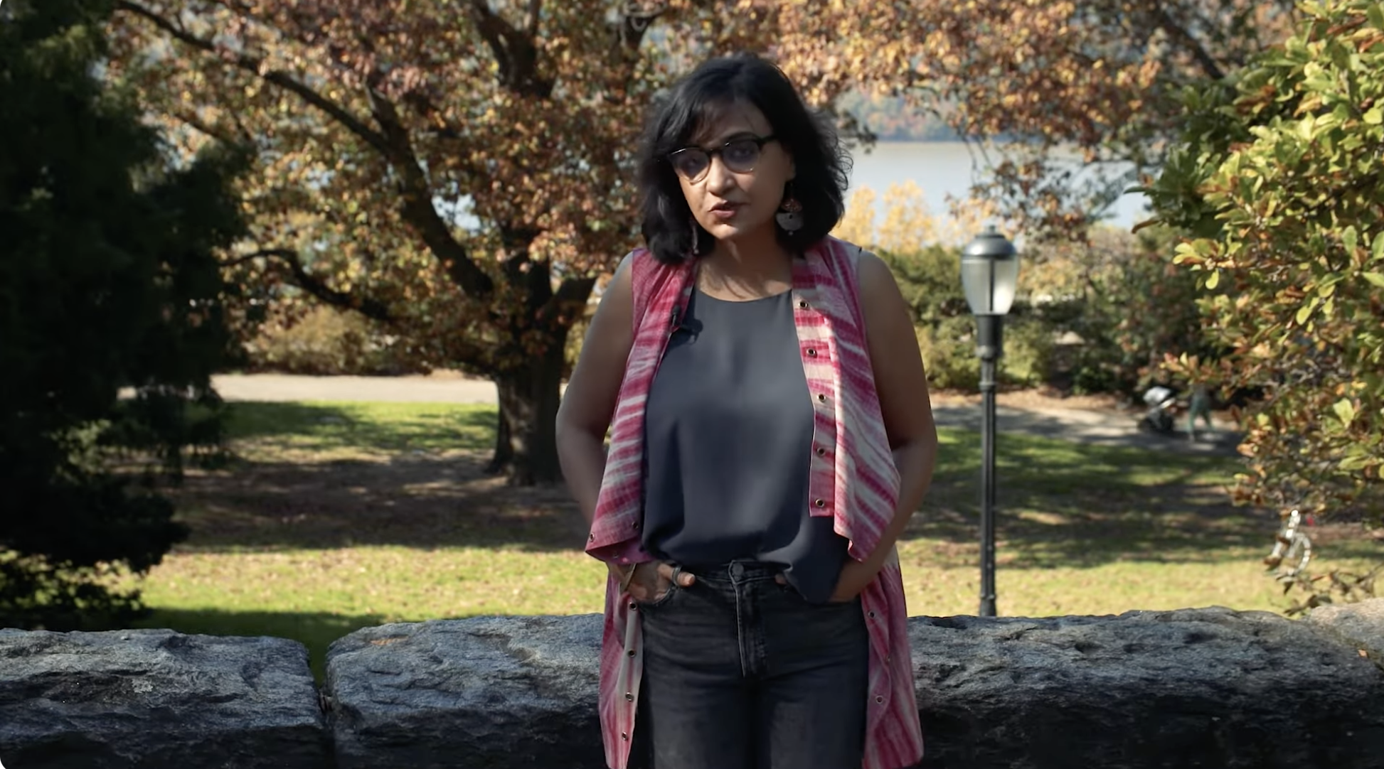 A woman with dark hair, glasses, and earrings standing outdoors near a stone wall, in a park with trees and autumn foliage, a lamppost, and a lake in the background.
