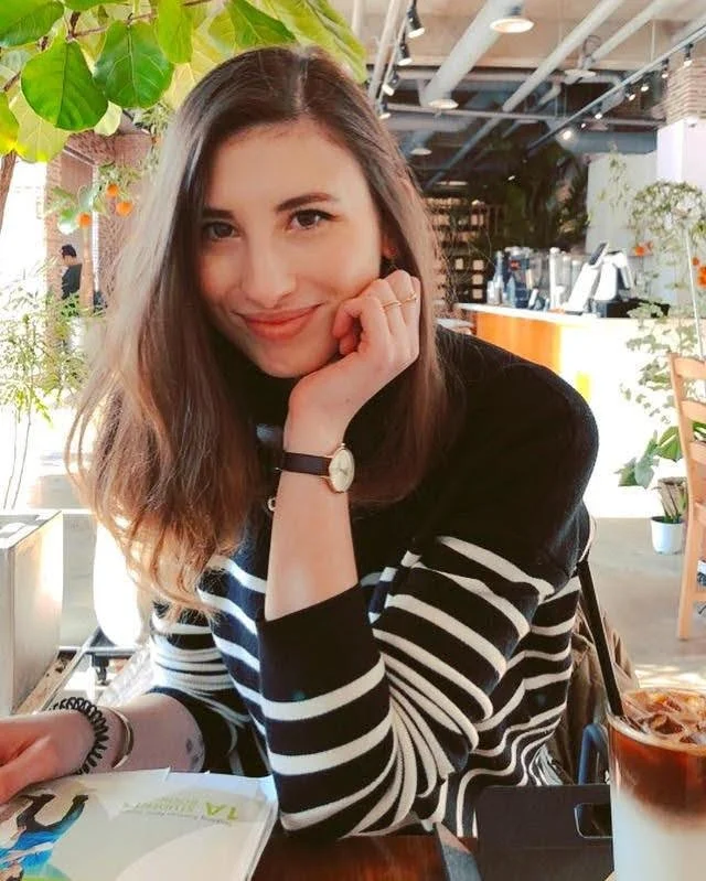 Une jeune femme souriante avec des cheveux bruns, portant un pull rayé noir et blanc, assise dans un café, avec des plantes vertes en arrière-plan.
