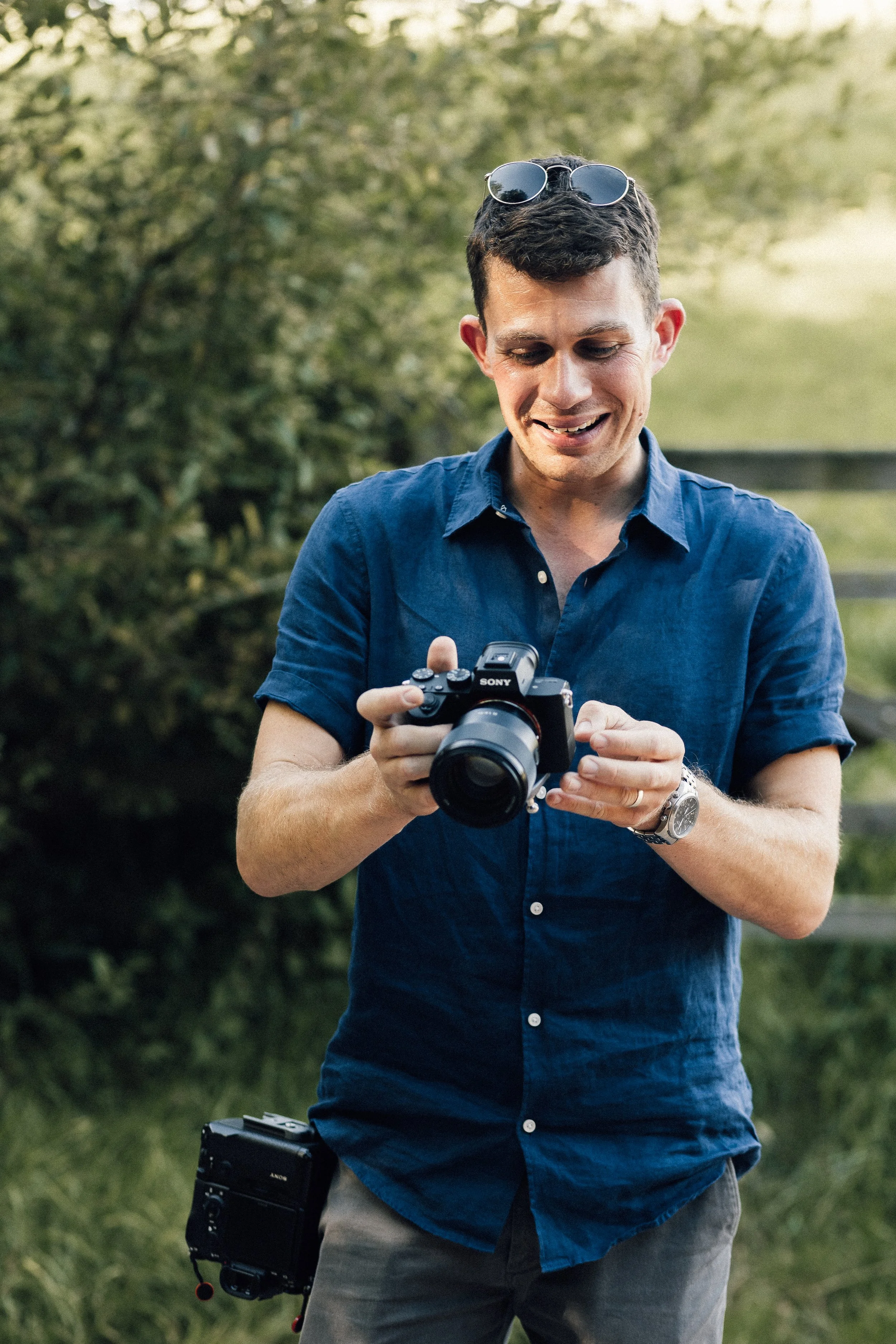 A man in a short-sleeved blue shirt and sunglasses resting on his head is outdoors, smiling while holding a Sony camera. There is greenery in the background.