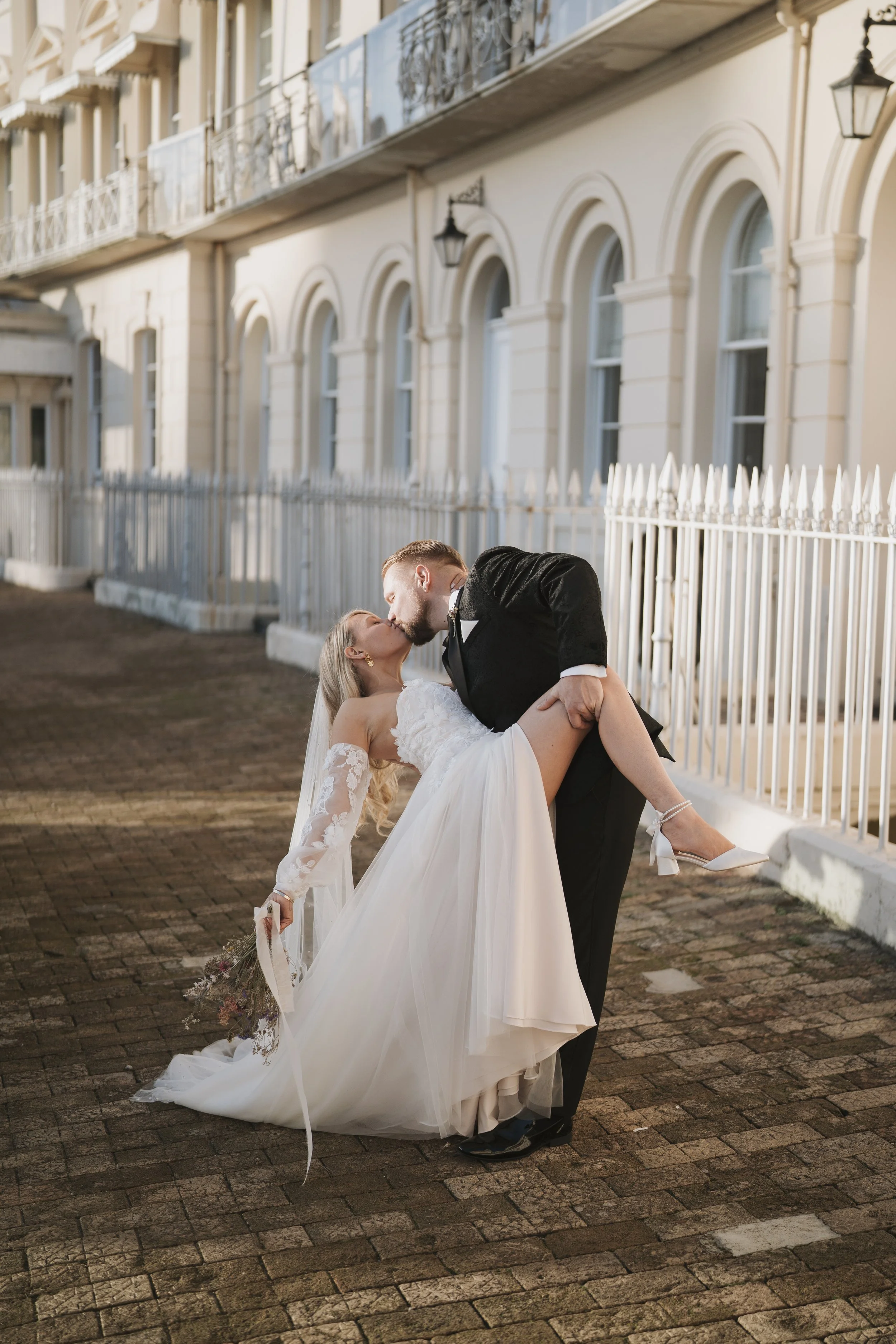A groom in a tuxedo carrying a bride in a wedding dress and lace sleeves, with a bouquet of flowers, outside on a cobblestone street, in front of a cream-colored building with white fence and black lanterns, during sunset.