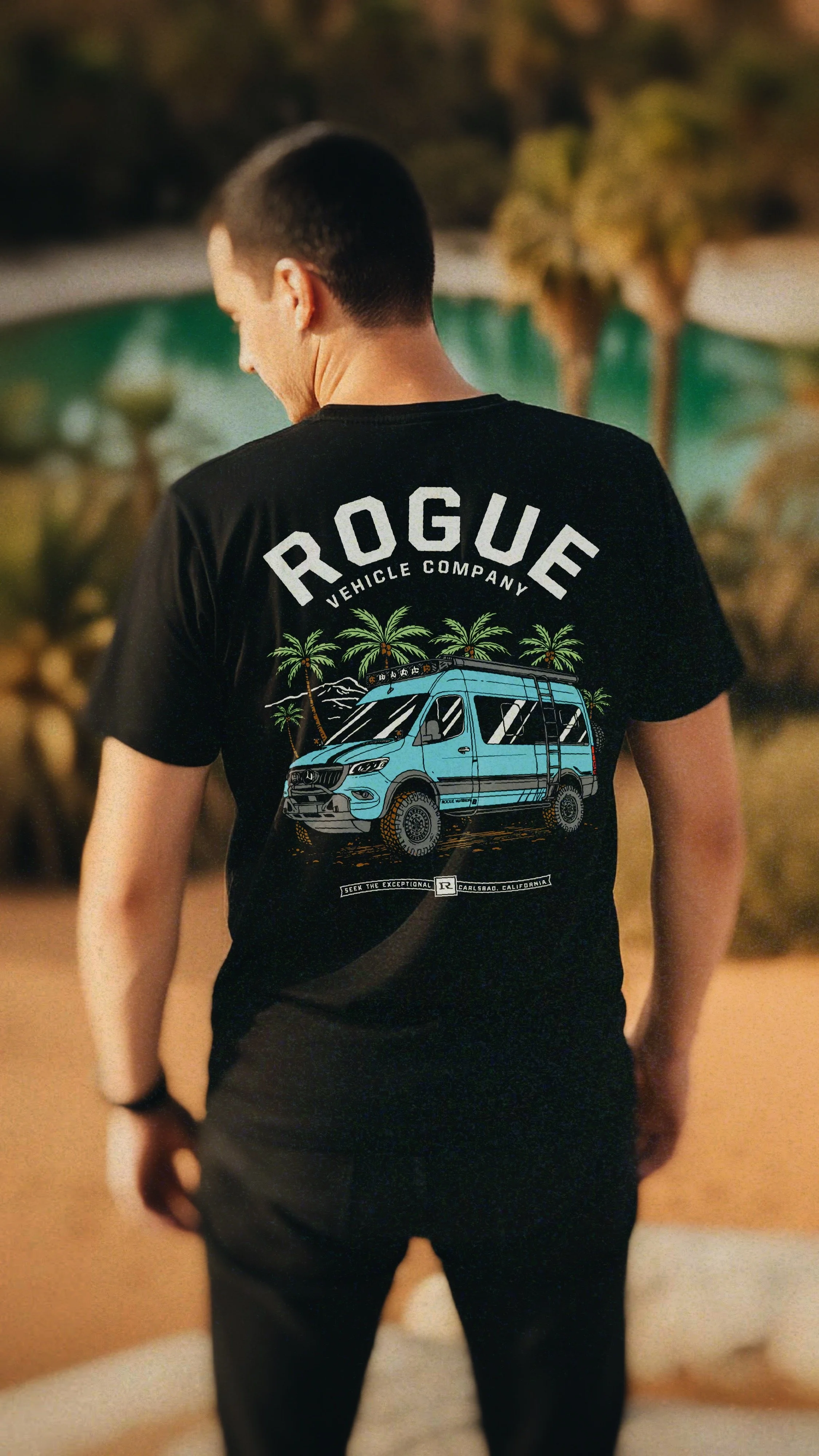 A person wearing a black T-shirt with a graphic of a blue van, palm trees, and mountains. The text on the shirt reads 'ROGUE Vehicle Company' and 'Seek the exceptional.' The person is standing outdoors, facing away, with their head turned slightly do
