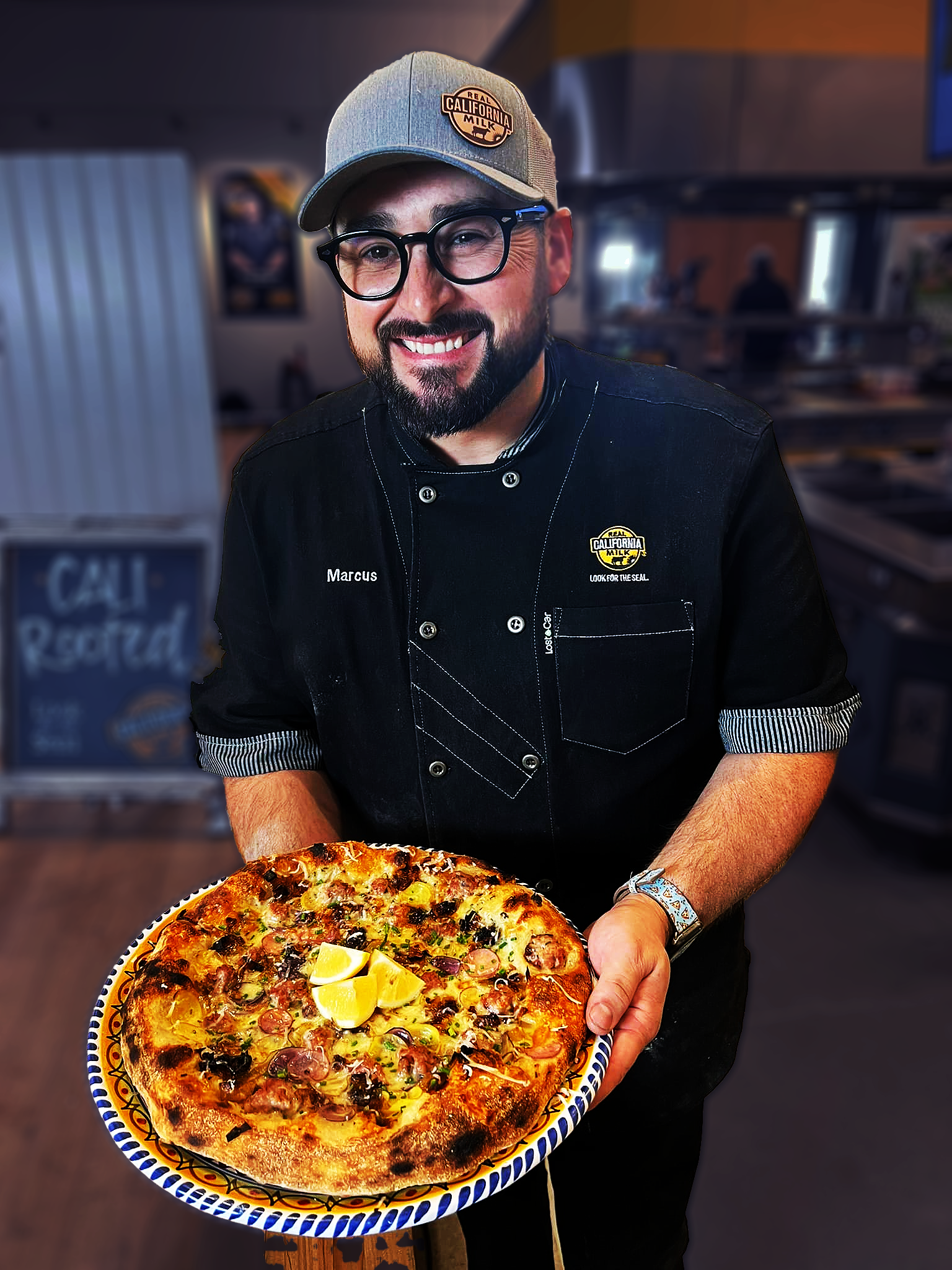 Man wearing glasses and a chef's jacket, holding a pizza with lemon slices on top in a restaurant kitchen.