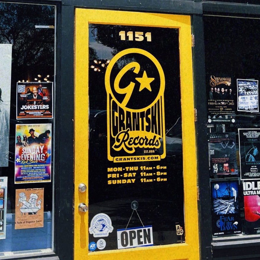 The entrance door of Grantski Records with a yellow and black logo. The store hours are Monday through Thursday, 11 am to 6 pm; Friday and Saturday, 11 am to 8 pm; Sunday, 11 am to 6 pm. There is an open sign on the door, and surrounding posters advertising events and music.