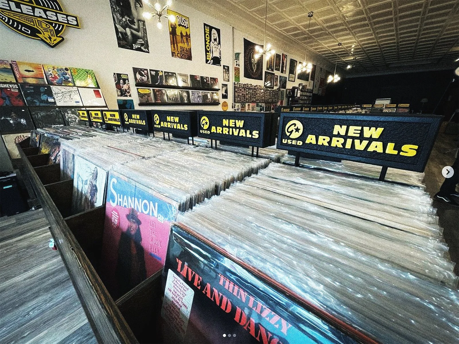 A record store with vinyl albums organized in bins and signs that read 'New Arrivals'. Posters and album covers decorate the walls.