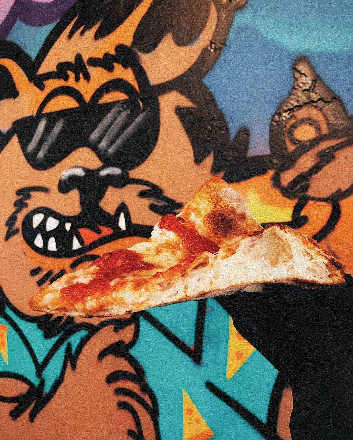 A hand wearing a black glove holding a slice of pizza with cheese and tomato sauce in front of a colorful graffiti mural featuring a cartoon animal character wearing sunglasses.