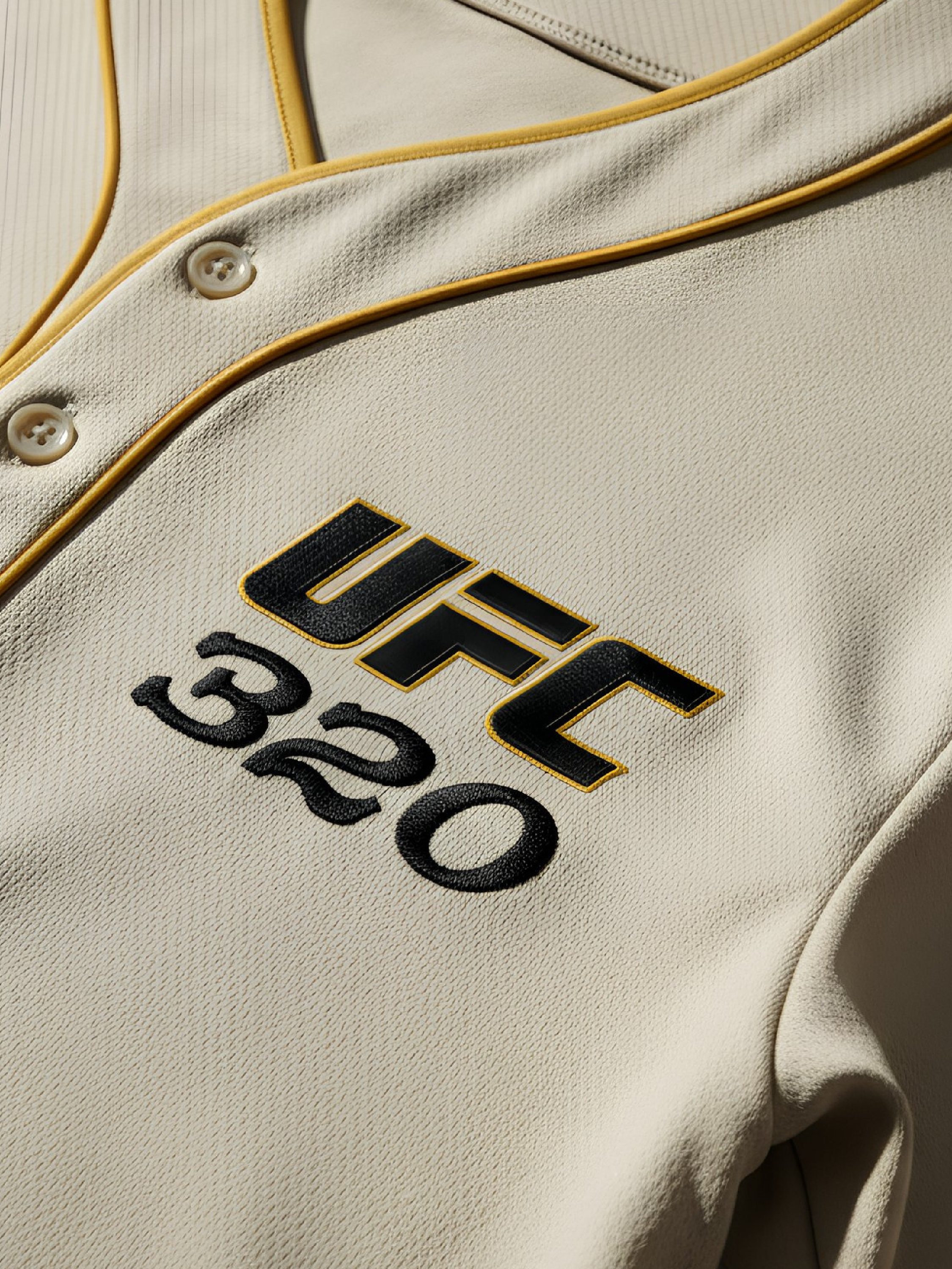 Close-up of a beige UFC jersey with black and gold embroidery, featuring buttons and a yellow piping detail.