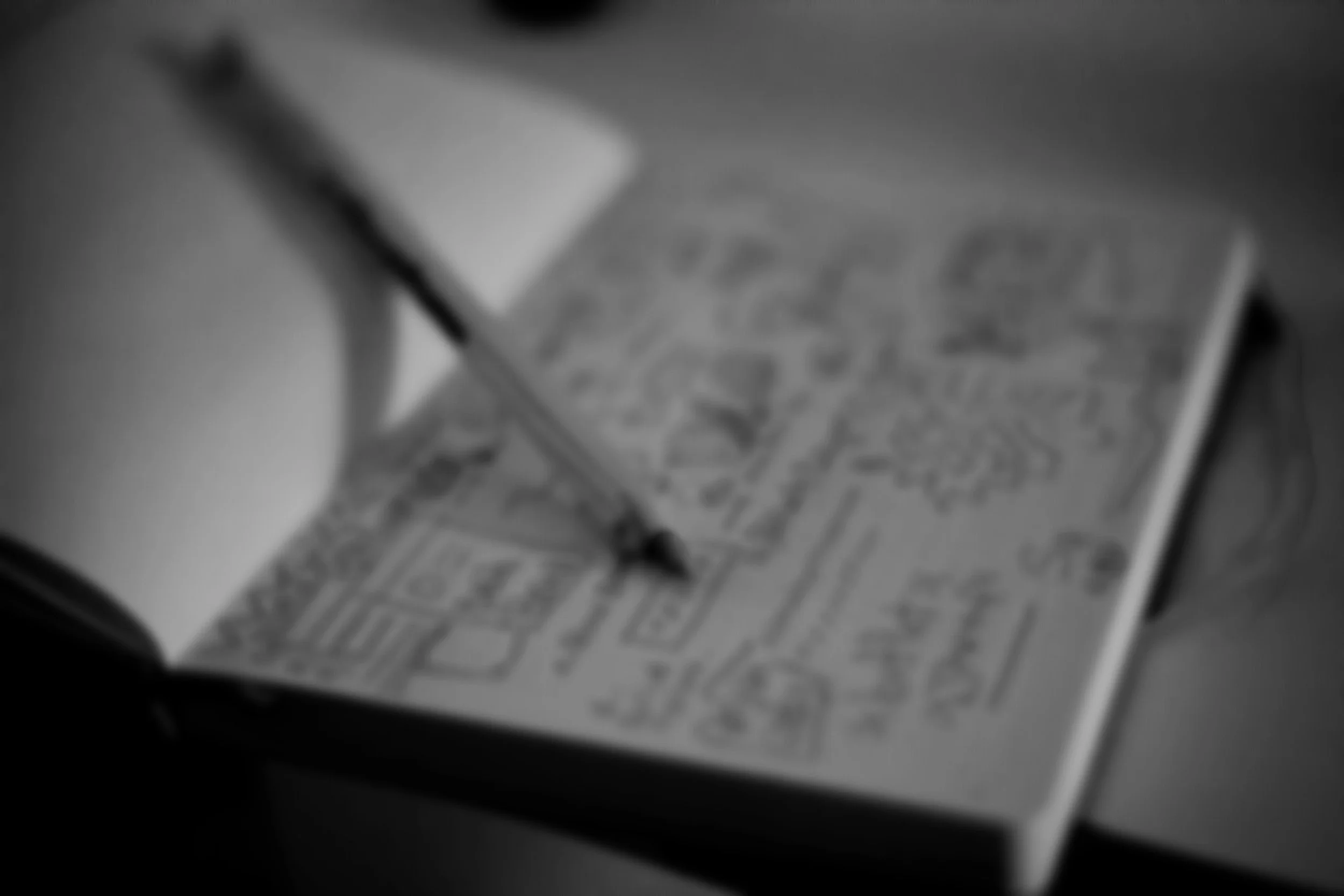 A blurred black and white image of an open coloring book with a pencil resting on it, lying on a flat surface.
