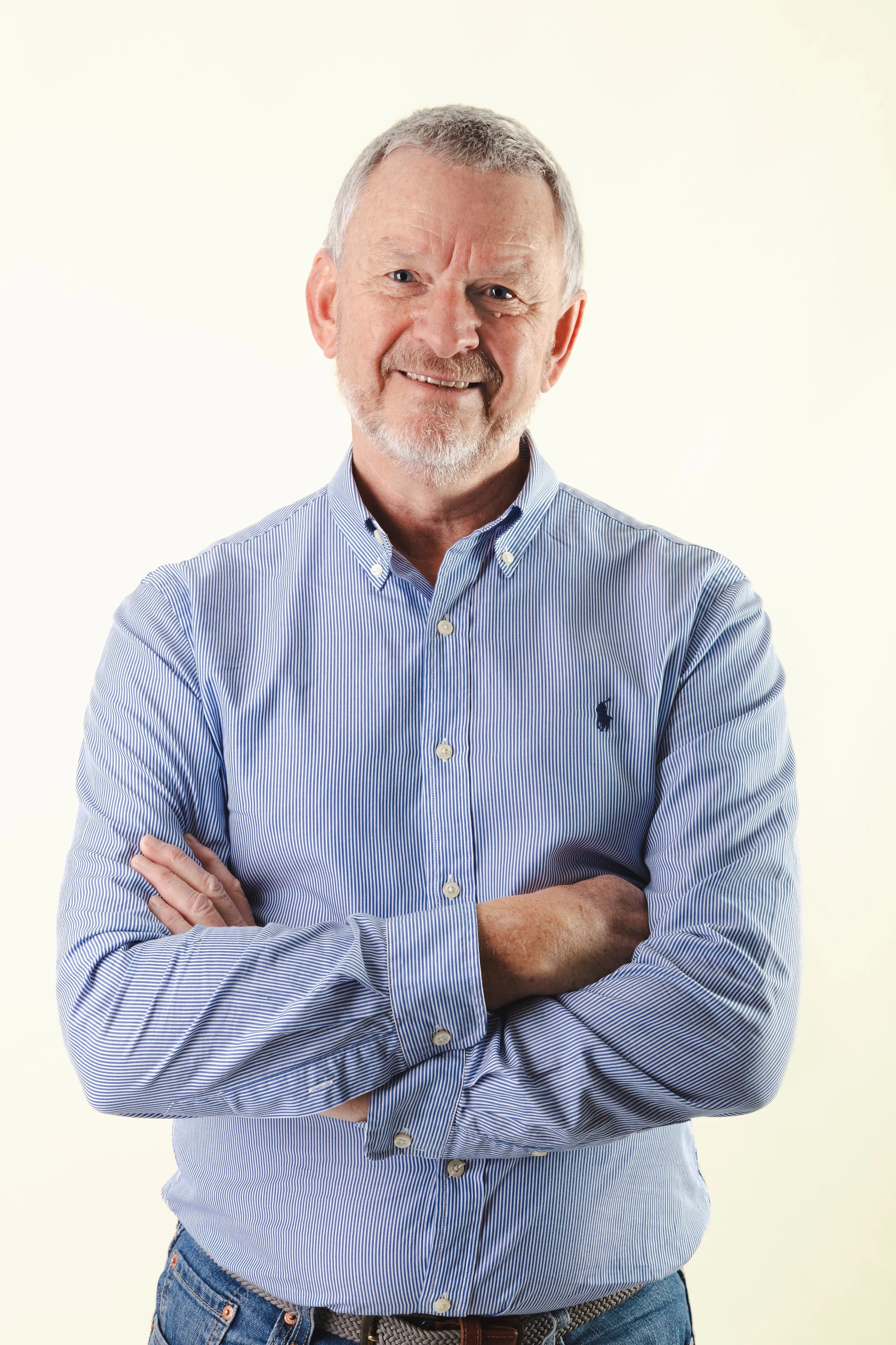 A middle-aged man with gray hair and beard smiling, wearing a blue striped button-up shirt with sleeves rolled up and crossed arms, standing against a plain, light background.