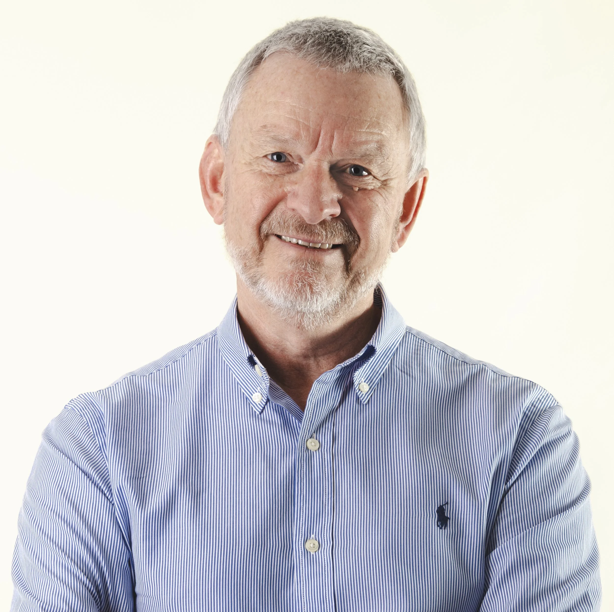 Close-up of an older man with gray hair, blue eyes, and a friendly smile, wearing a blue and white striped dress shirt against a plain light background.