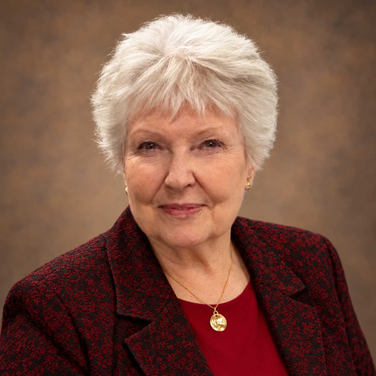 An elderly woman with short white hair and light skin, wearing a dark red blouse, a black and red patterned blazer, gold earrings, and a gold necklace with a round pendant. She is smiling gently against a blurred brown background.