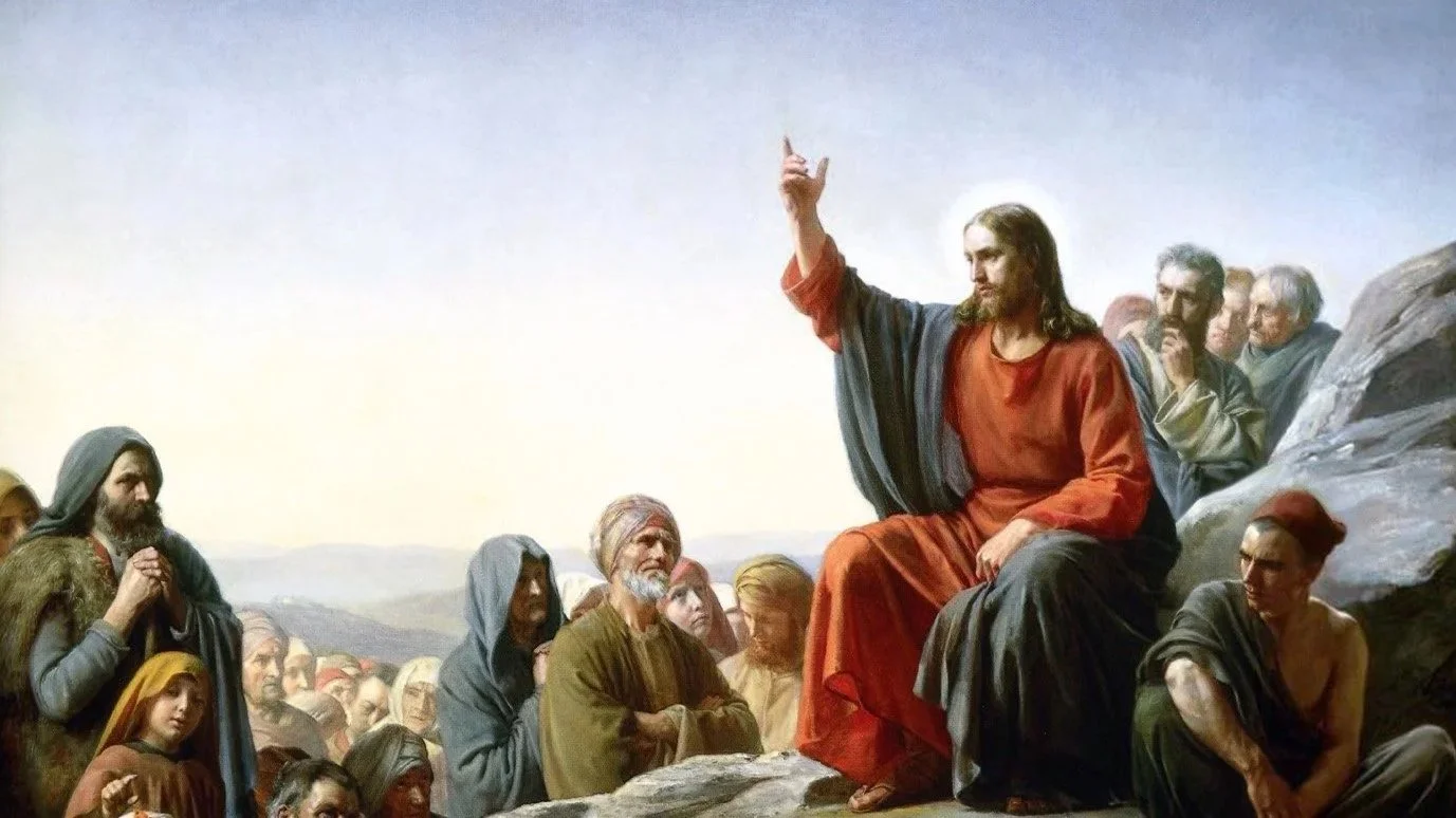 Jesus, vested in red and dark blue, sits on a rocky outcropping with his right hand raised in a teaching gesture. A varied crowd surrounds him—disciples, women, children, the elderly, the poor—gathered on the hillside against a dawn-lit landscape.