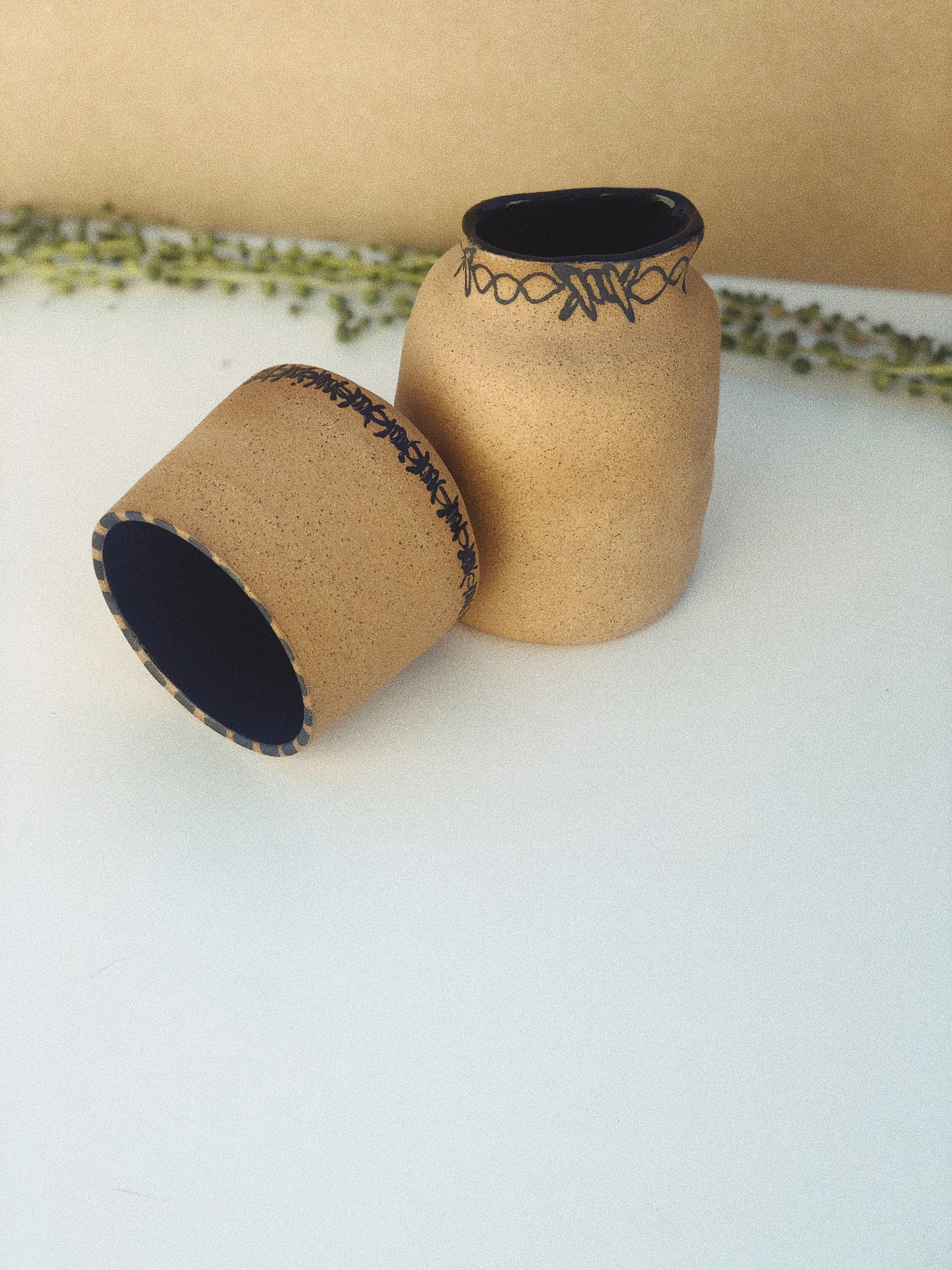Two small beige pots with black interior and decorated rims, one placed upright and the other lying on its side, on a white surface with a beige background and some greenery in the background.