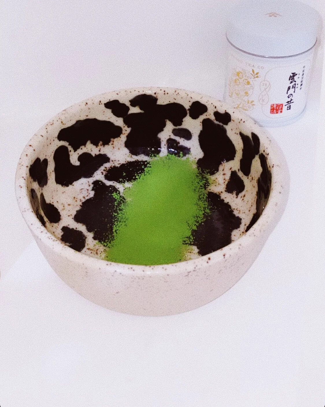 A bowl with a black and white cow pattern interior and bright green matcha powder in the center. A container with Japanese writing is in the background.