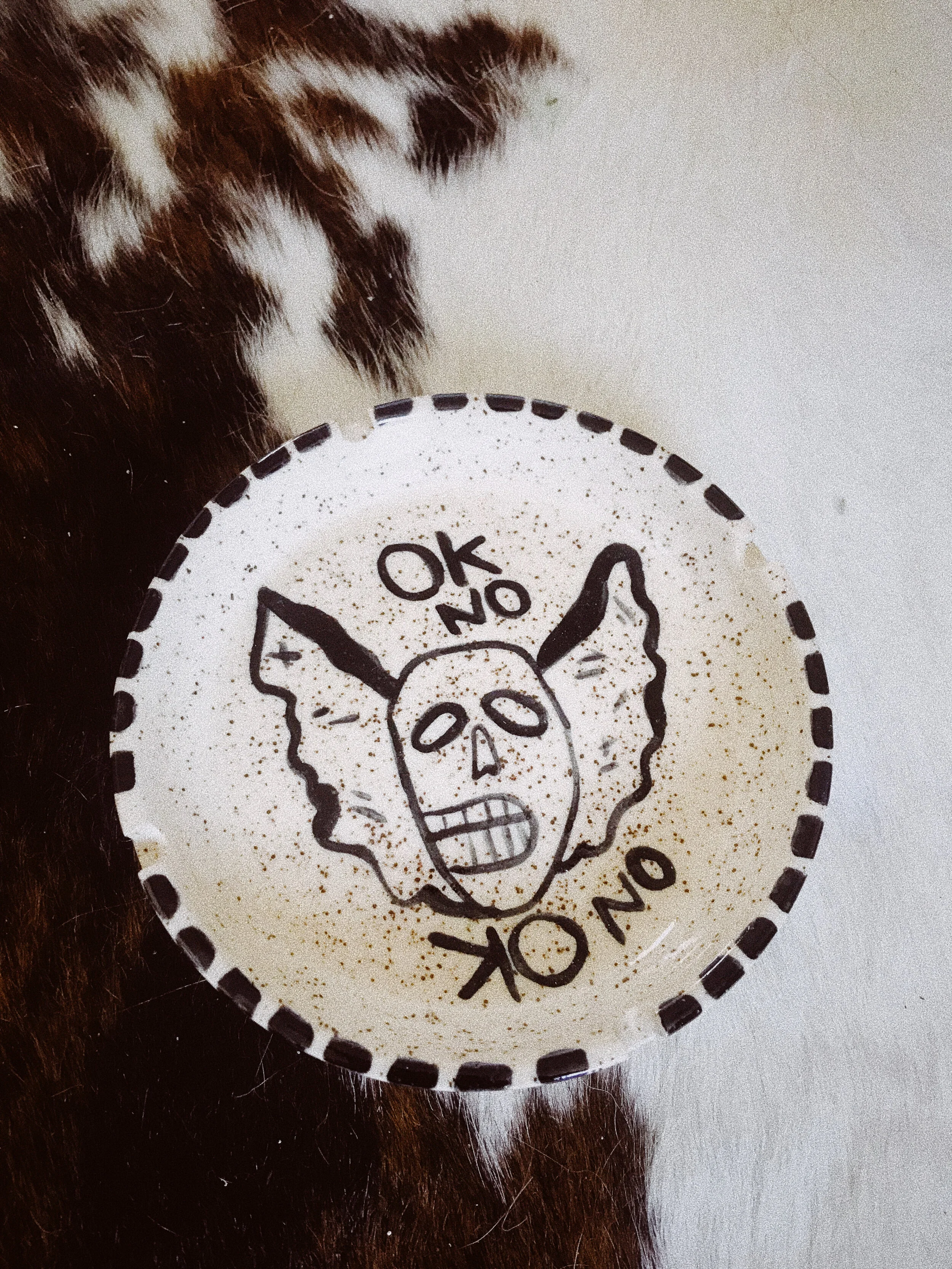 A paper cup with a drawing of a skull wearing bat wings, with the words 'OK NO' written above and below the skull, and a black dashed border.