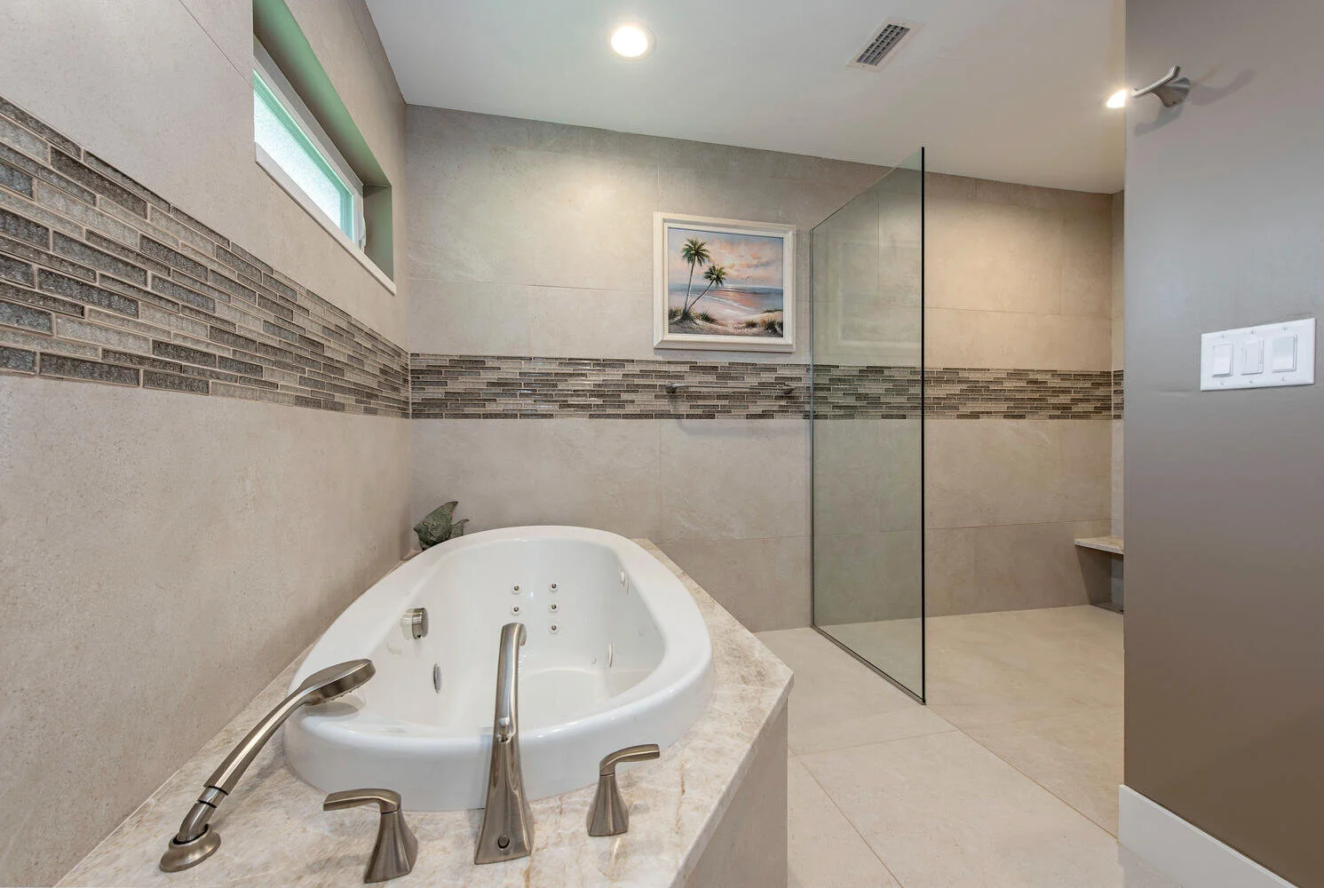 Modern bathroom with a whirlpool tub, glass shower enclosure, small window, framed beach scene painting, and neutral-colored tiles and walls.