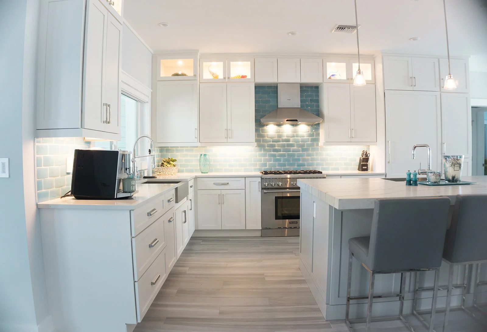 Bright modern kitchen with white cabinets, light blue subway tile backsplash, stainless steel stove and range hood, kitchen island with seating, and pendant lighting.