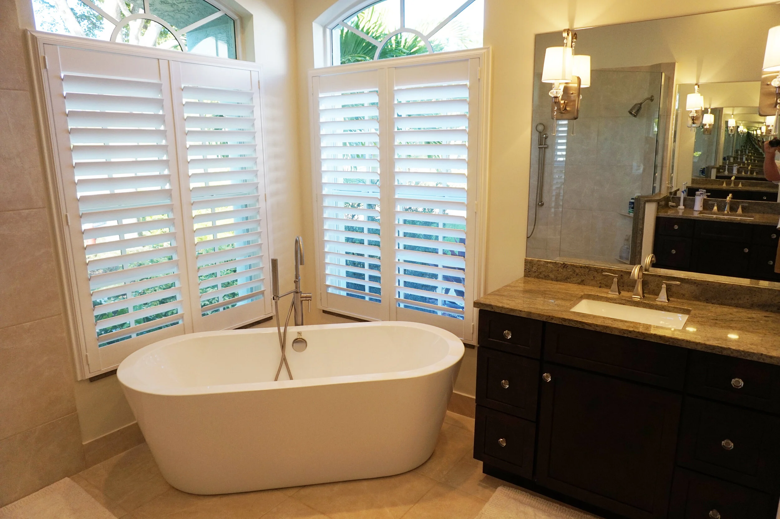 A bathroom with a freestanding bathtub positioned in front of large windows with white plantation shutters. To the right, there is a dark wooden vanity with a granite countertop, a sink, and a mirror. There are wall-mounted light fixtures above the v