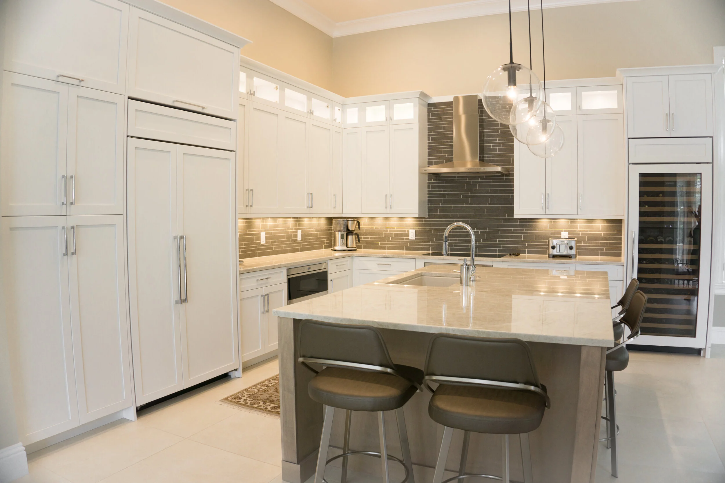 Modern kitchen with white cabinets, beige countertops, a gray tile backsplash, and pendant lighting above a kitchen island. There are chairs around the island, a built-in microwave, and a wine fridge on the right.