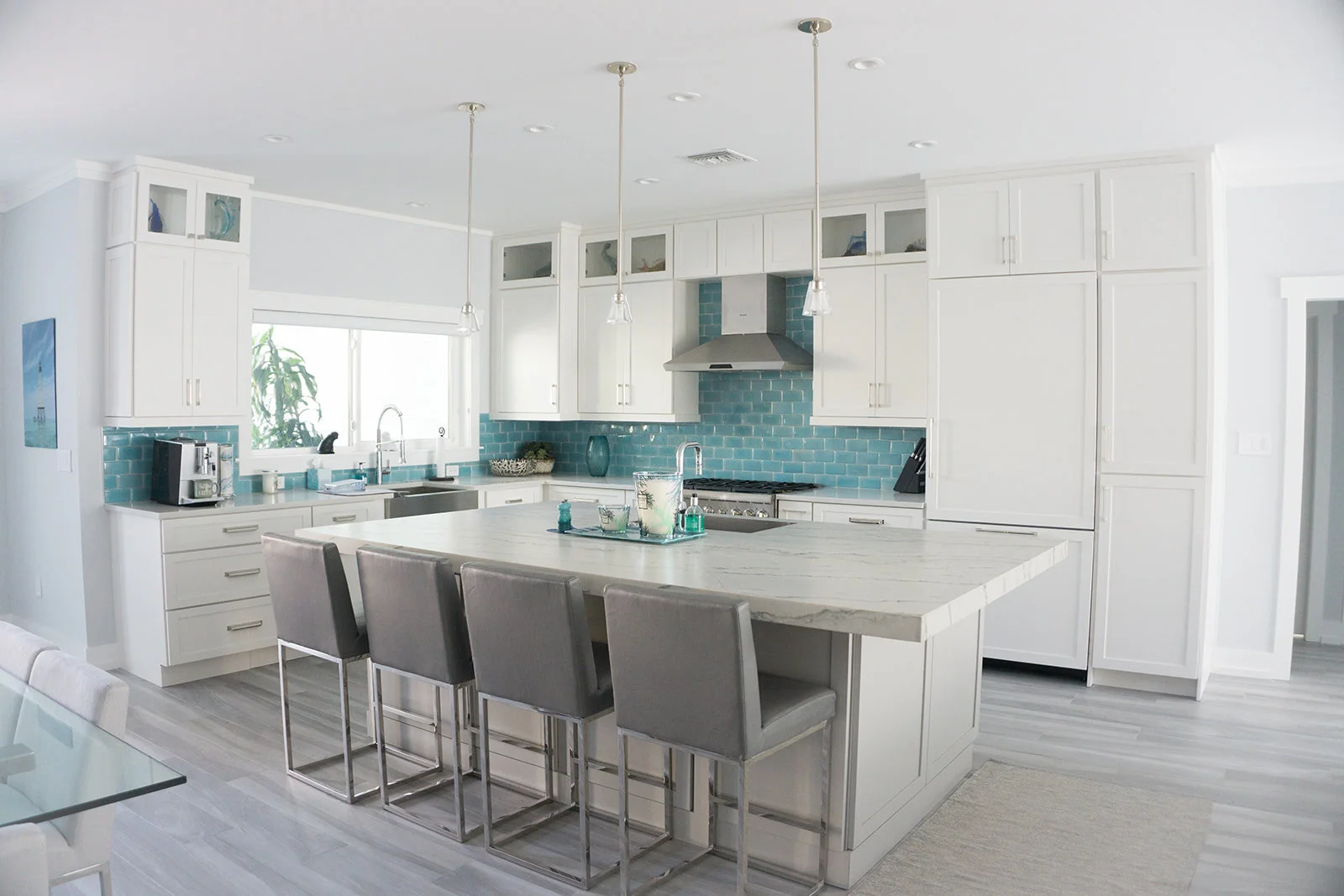 Modern white kitchen with a large island, gray chairs, blue tile backsplash, stainless steel appliances, and a window overlooking greenery.