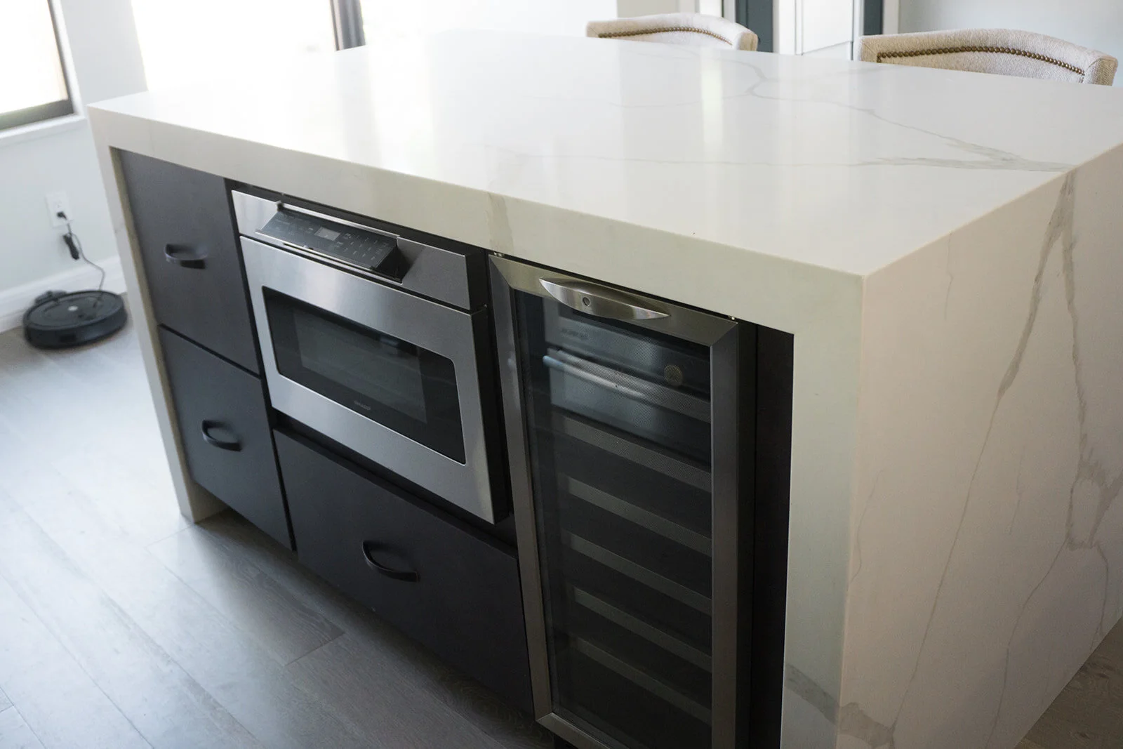 A kitchen island with a white marble countertop and dark gray base, featuring a built-in microwave oven, a wine cooler with glass door, and storage drawers.