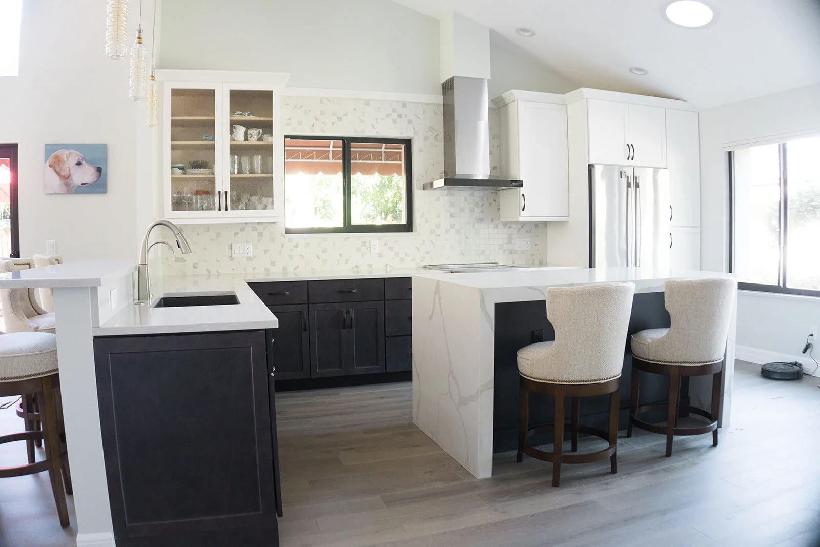 Modern kitchen with black and white cabinetry, marble island, and beige bar stools, large windows, and a light-colored hardwood floor.