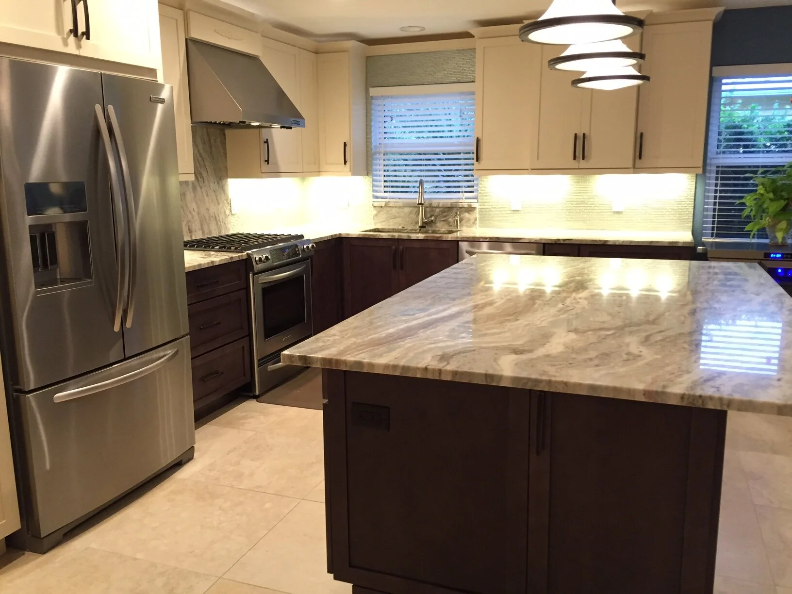 Modern kitchen with a large marble kitchen island, stainless steel refrigerator, oven, range hood, upper and lower cabinets, a window with blinds, and pendant lighting.