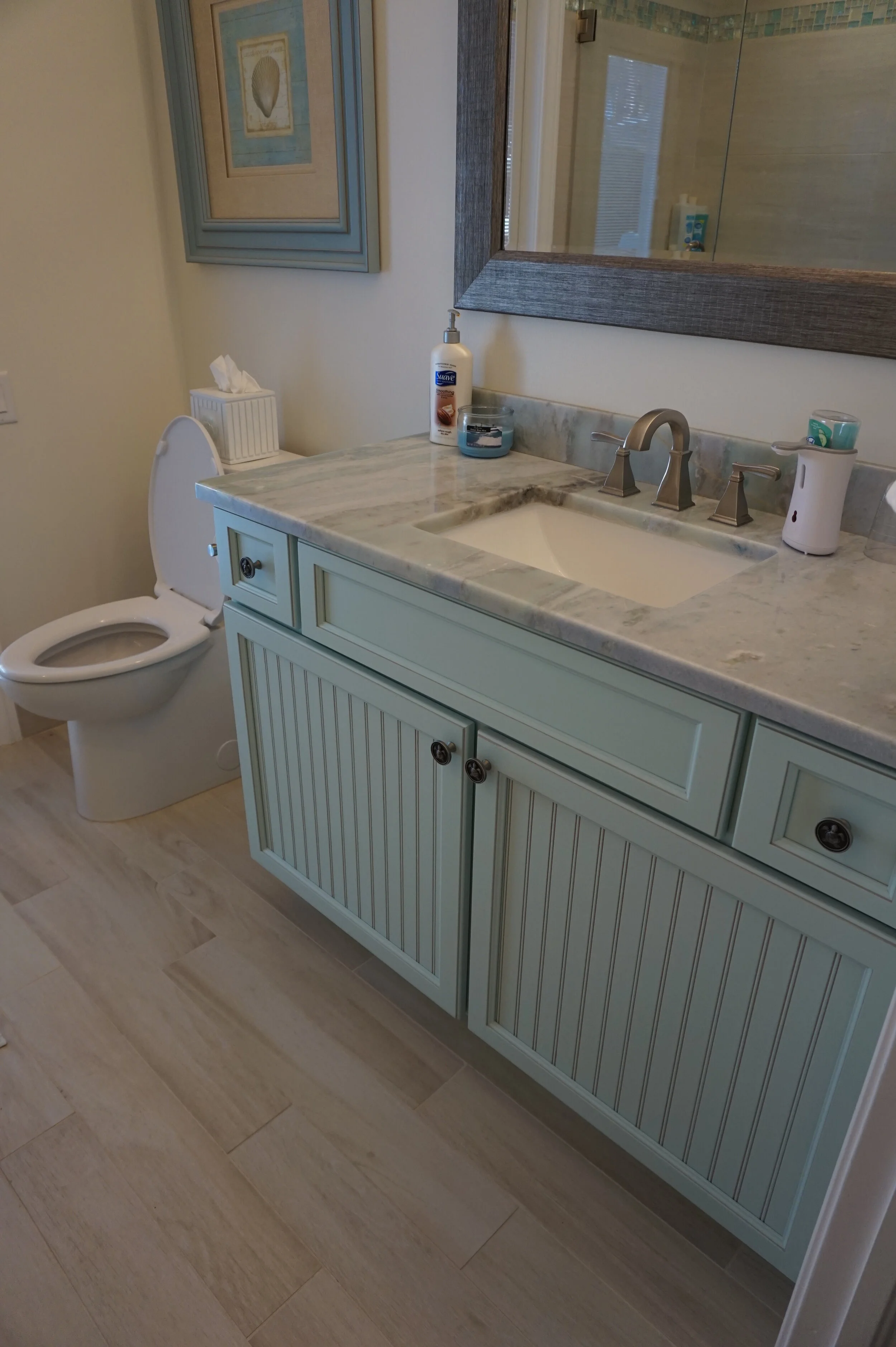 A bathroom with a toilet and a light green vanity with a marble countertop. The vanity has a large mirror above it and various toiletries on the countertop. There is a framed picture of a shell hanging on the wall.