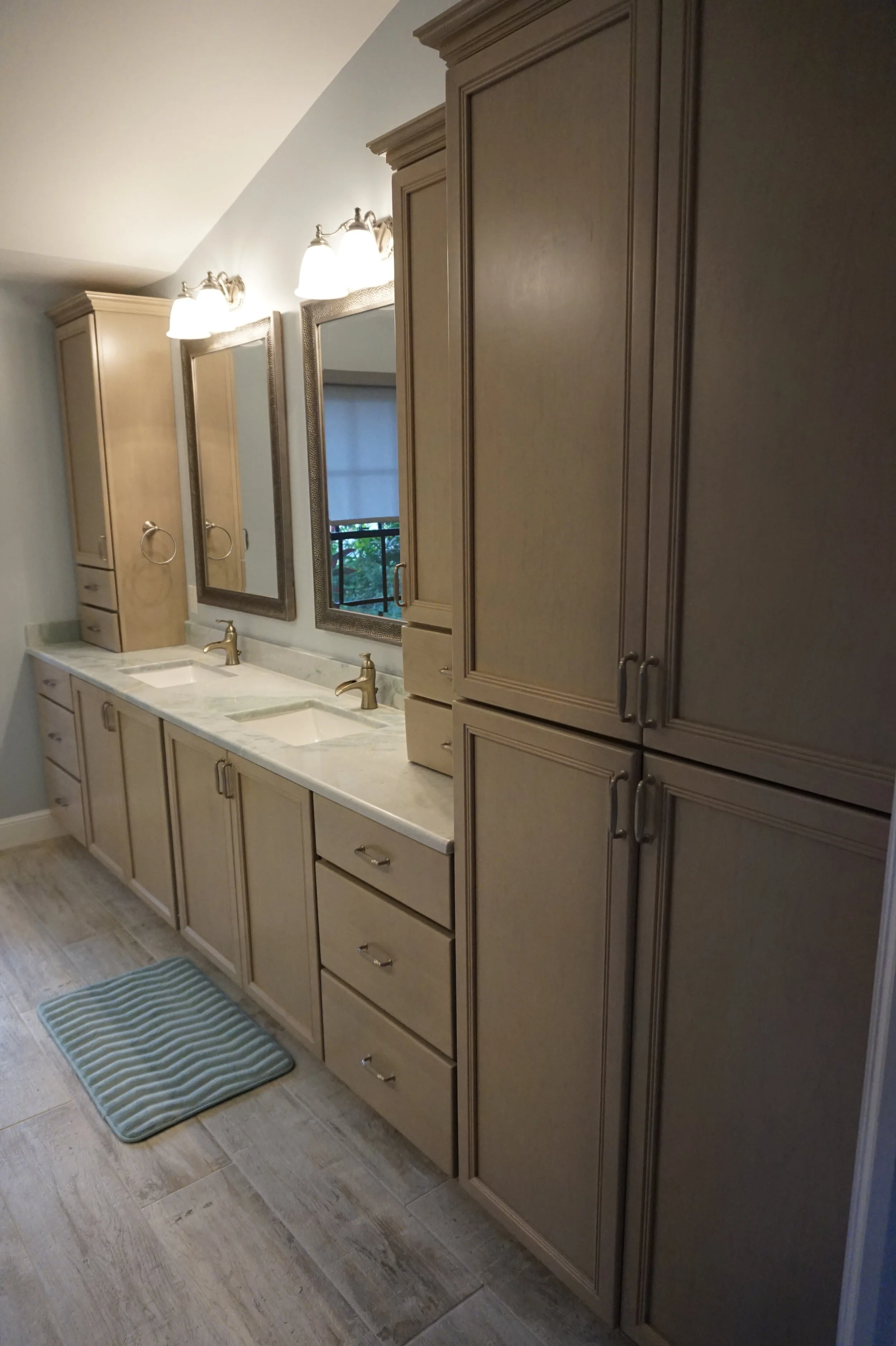 Bathroom with beige cabinets, a marble countertop, two sinks, and two mirrors. Three light fixtures are mounted above the mirrors. A window with a view of greenery is visible in the background. There is a striped bathmat on the tiled floor.