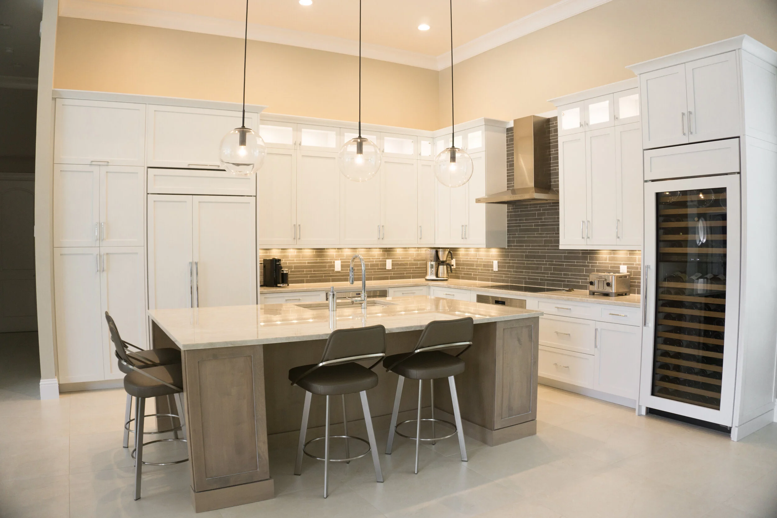 Modern kitchen with white cabinets, a kitchen island with gray chairs, black pendant lights, microwave, coffee maker, toaster, and wine cooler.