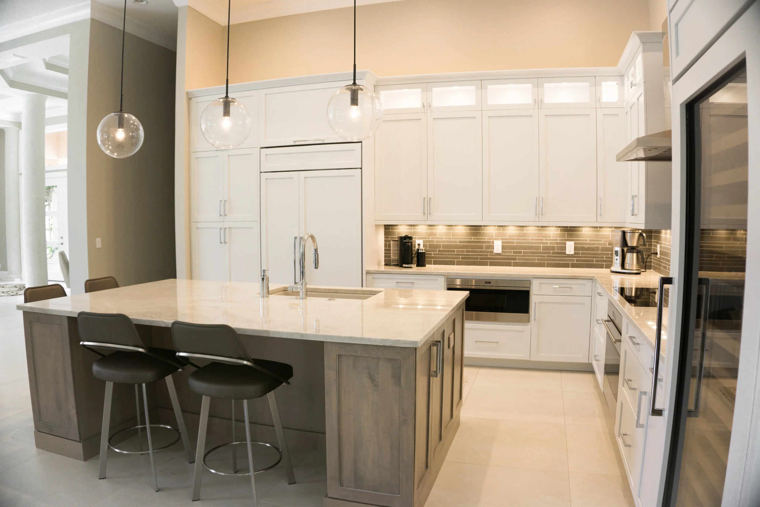 Modern kitchen with white cabinetry, a large marble island with four black stools, pendant lights, a dark brick backsplash, and stainless-steel appliances.