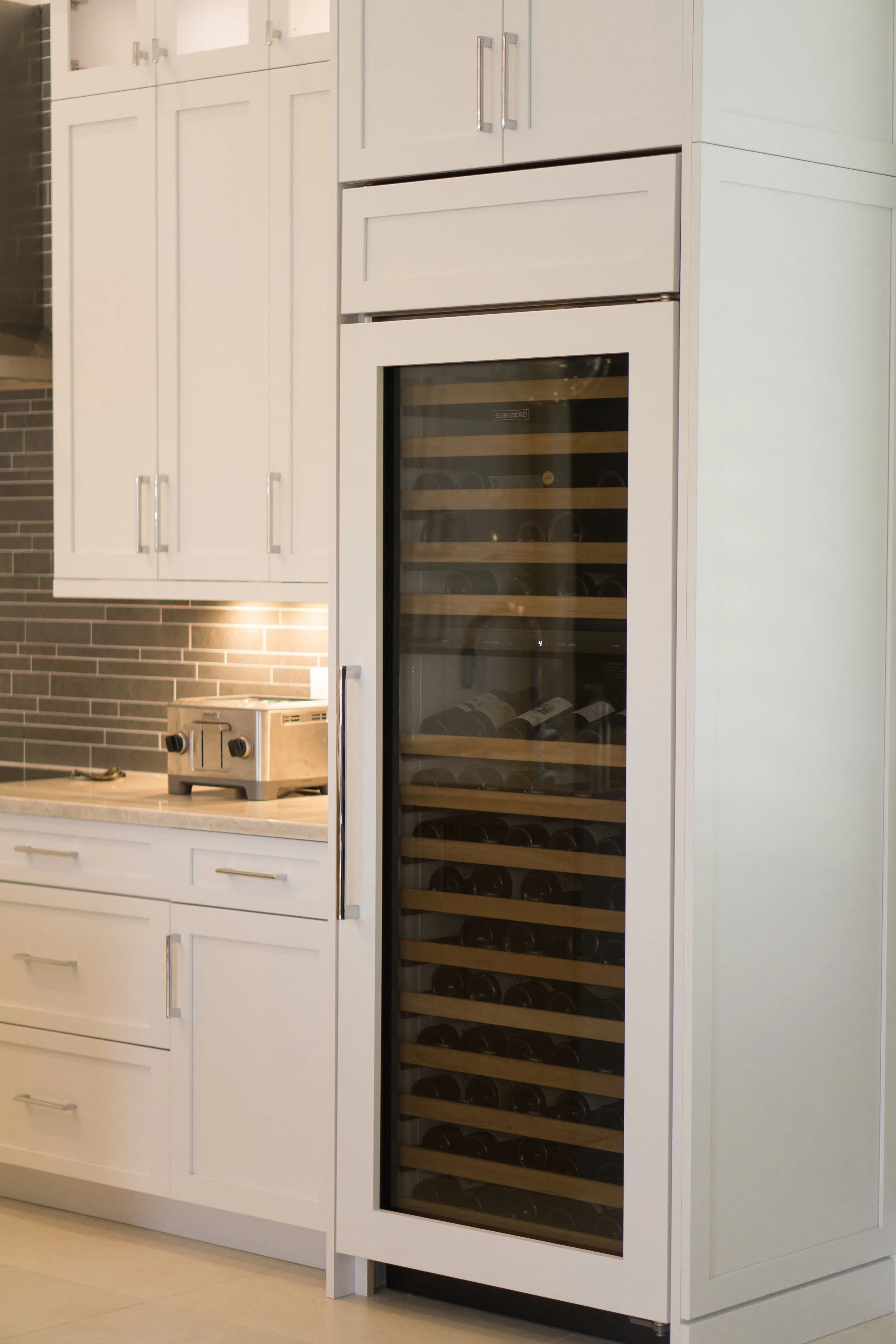 A white built-in kitchen cabinet with a wine cooler with a glass door, and a toaster on the counter below. The wine cooler contains several bottles stacked horizontally.