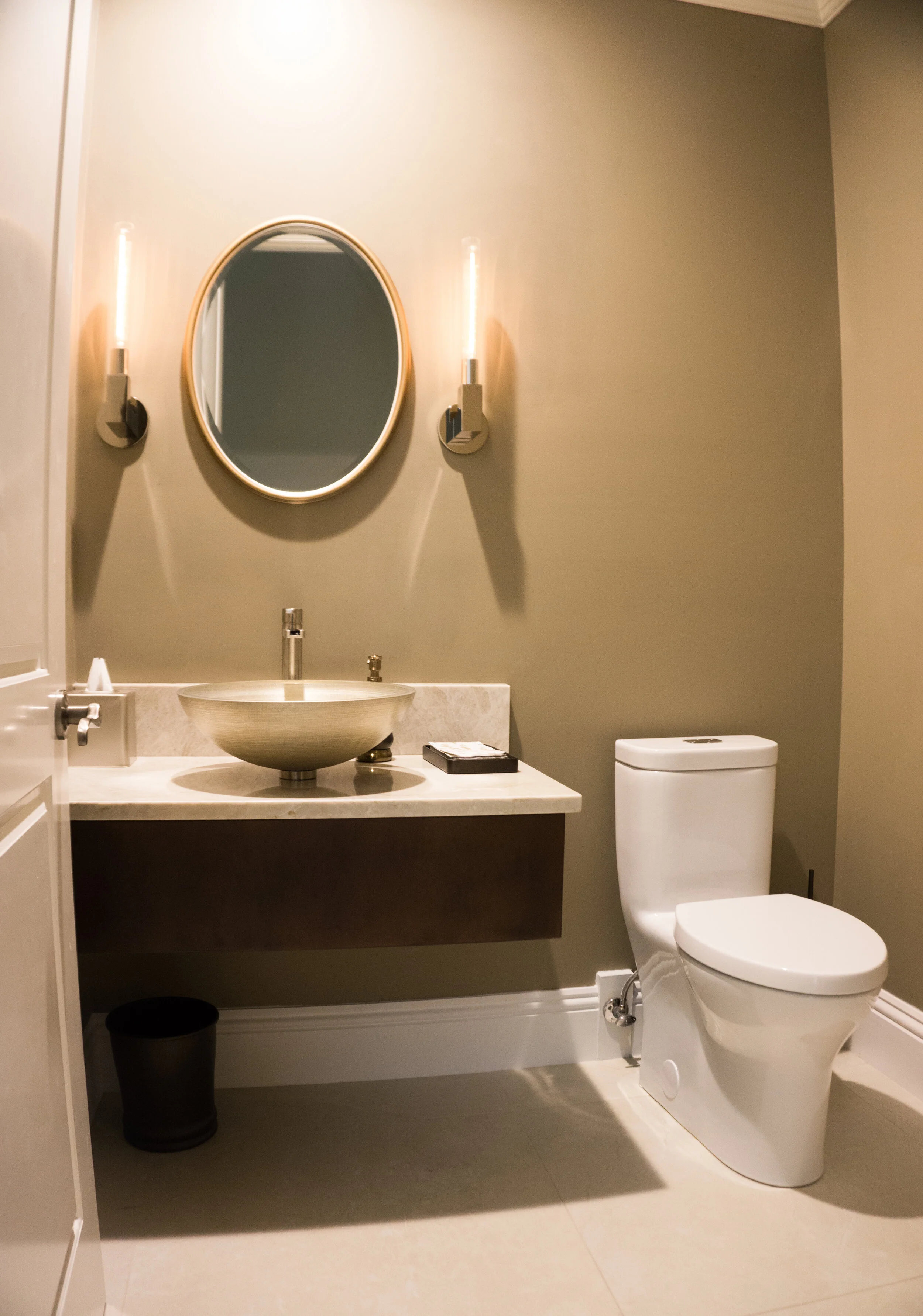 Modern bathroom with a wall-mounted mirror, two vertical wall lights, a vessel sink with a faucet, a soap dispenser, a small black box, and a toilet, with a small black waste bin below the counter.