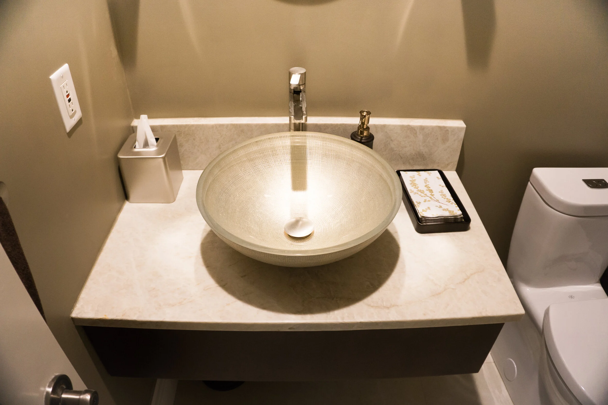 A bathroom vanity with a modern round glass vessel sink, a tall chrome faucet, a soap dispenser, a decorative tray, and a tissue box. A toilet is partially visible to the right.