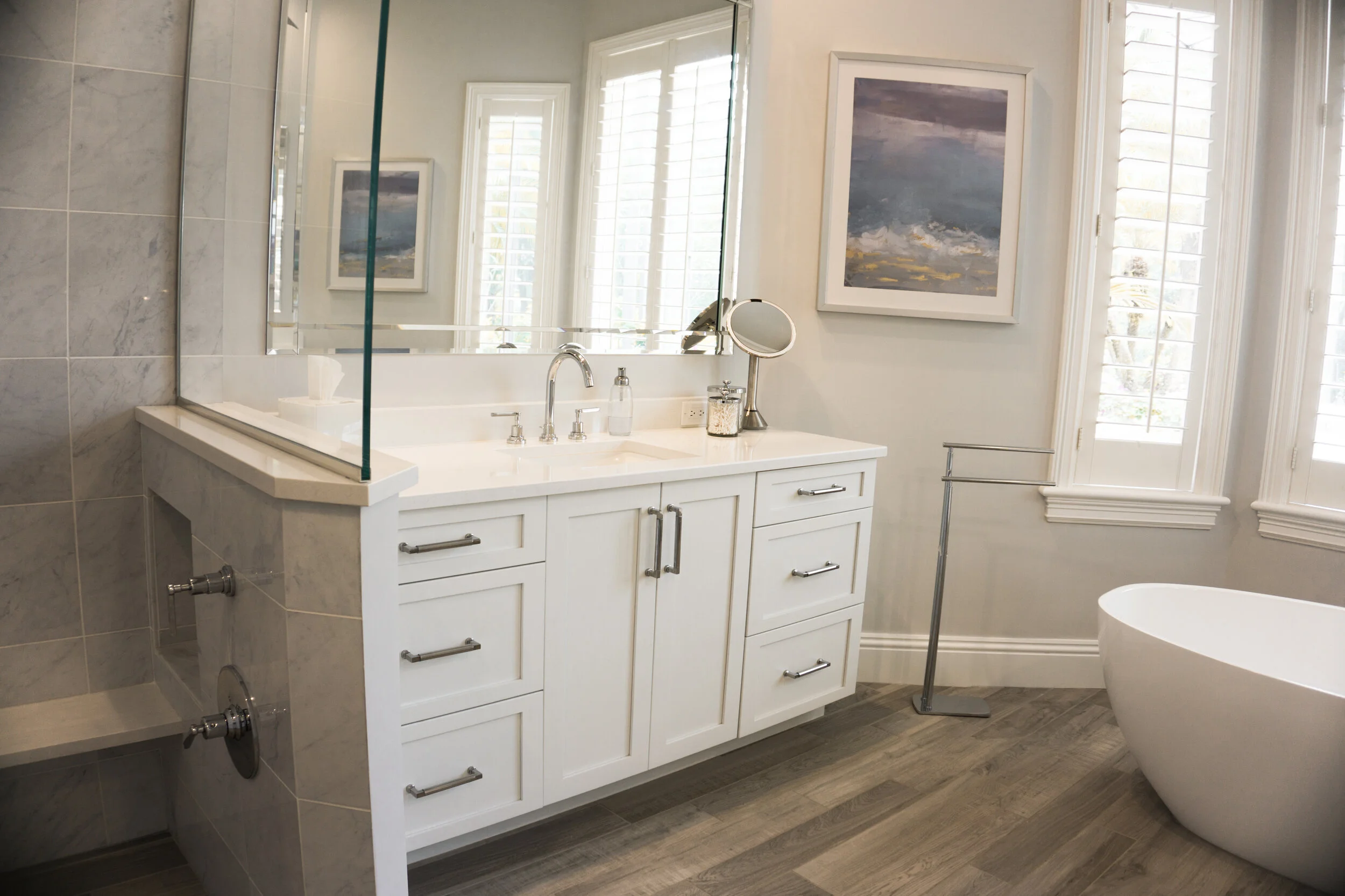 A modern bathroom with a white vanity cabinet, stainless steel handles, and a white countertop featuring a faucet, a soap dispenser, and a jar of cotton swabs. Above the vanity is a large mirror reflecting windows with white plantation shutters. To t