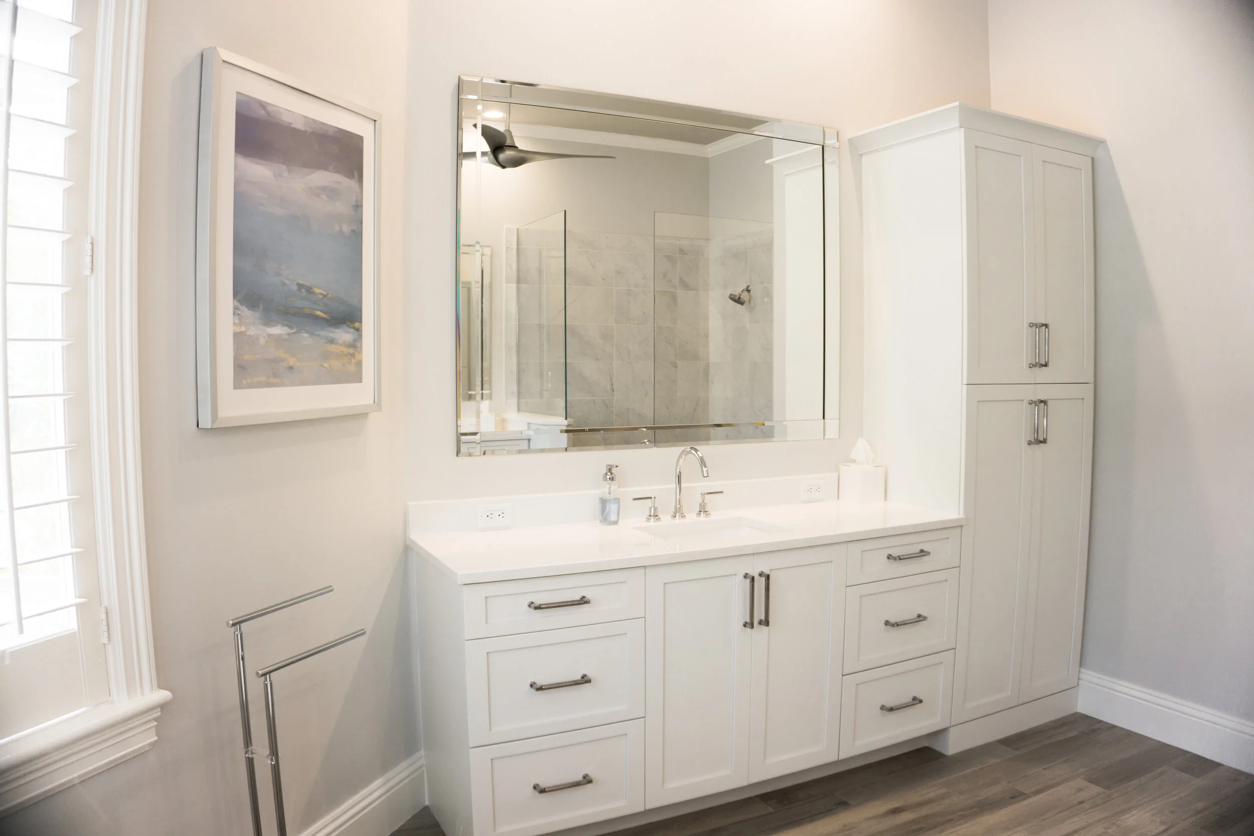 A white bathroom vanity with multiple drawers and cabinets, a double sink with silver faucets, a large wall mirror, a white tissue box, and a drying rack. To the right, there is a tall white cabinet. A painting of an abstract ocean scene is on the le