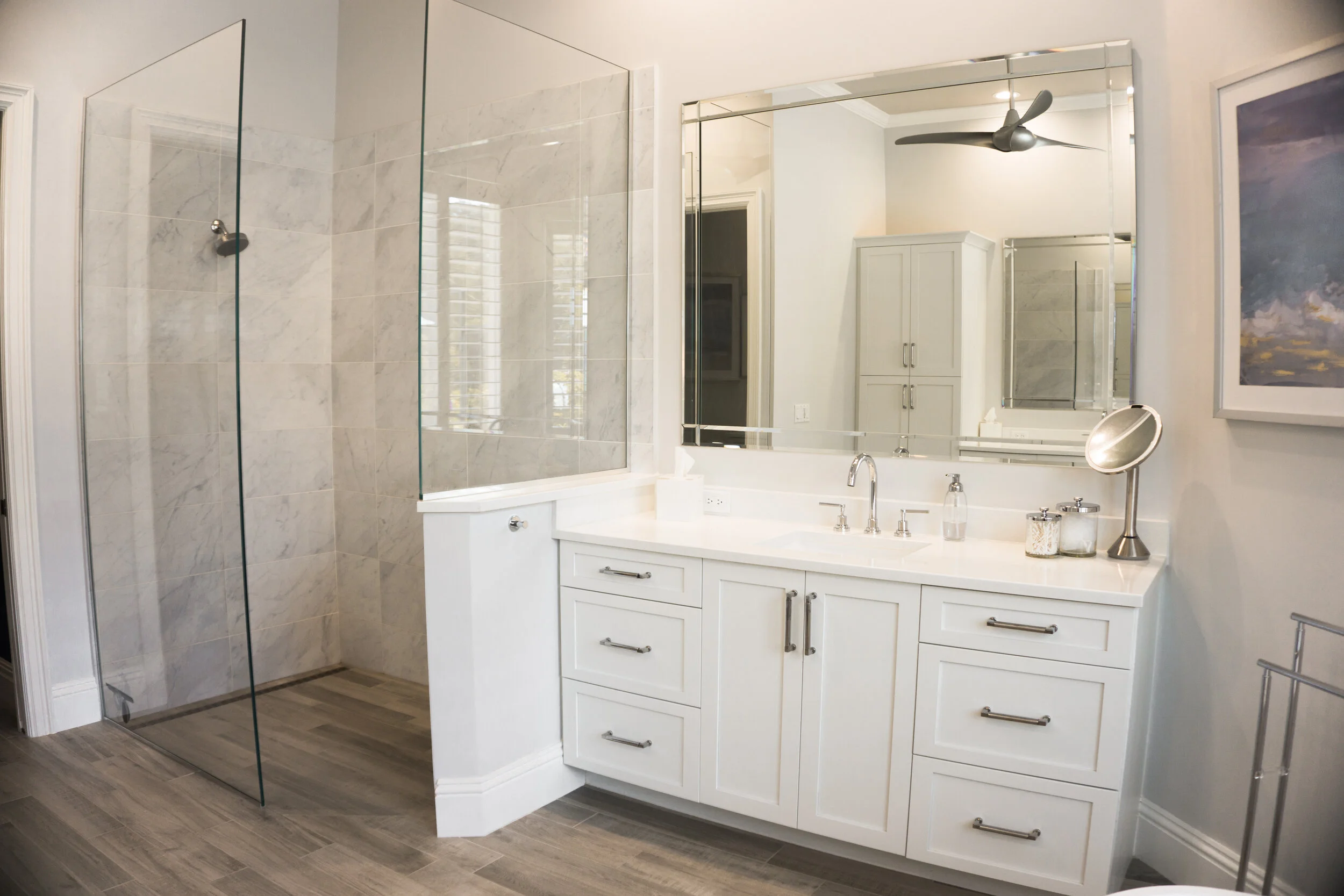 A modern bathroom with a glass-enclosed shower, a white vanity with a large mirror, and a ceiling fan. The room features white walls, a framed artwork, and wood-look tile flooring.