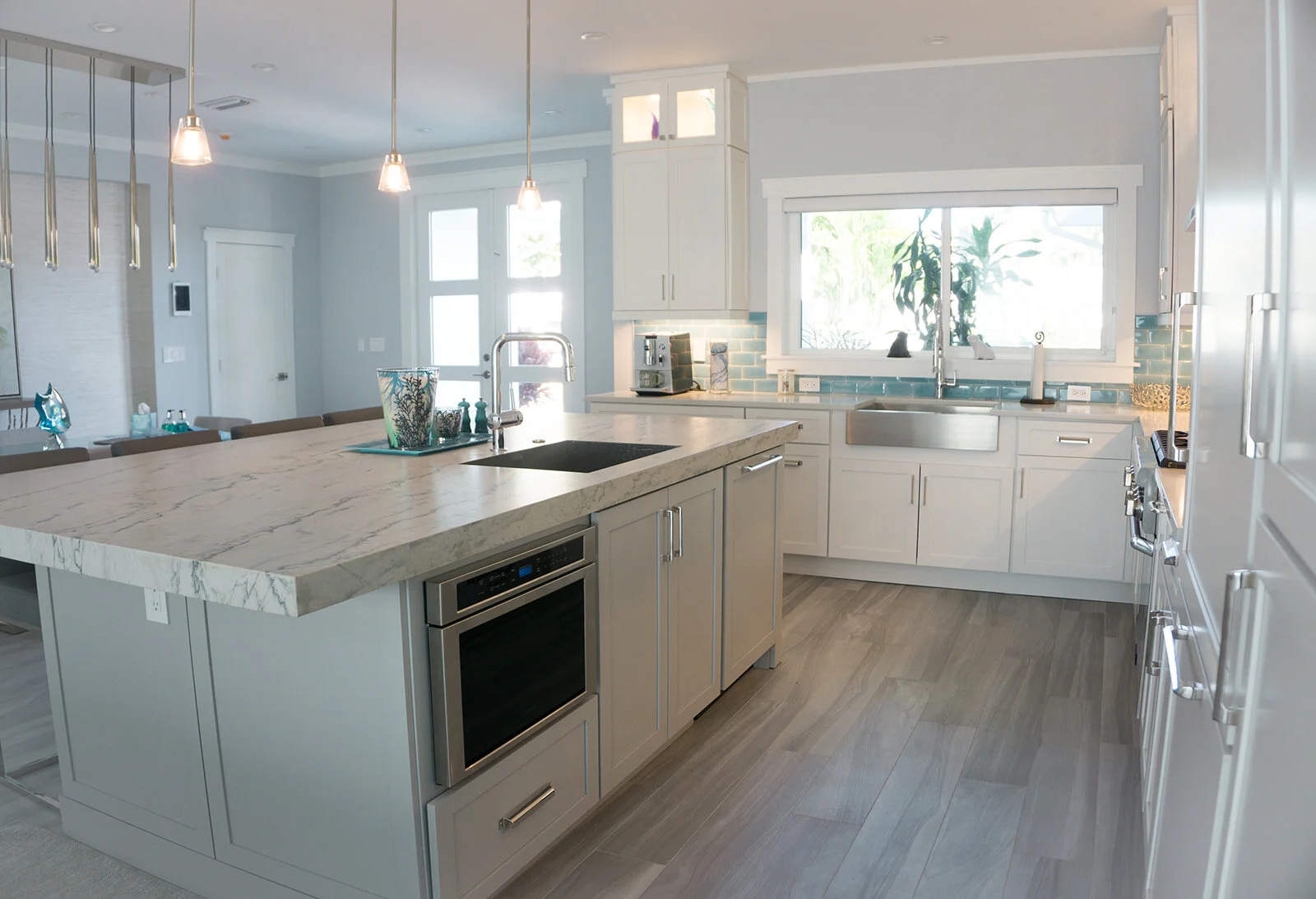 Modern kitchen with white cabinetry, marble island, stainless steel appliances, and light wood flooring. Bright window above the sink and pendant lights overhead.