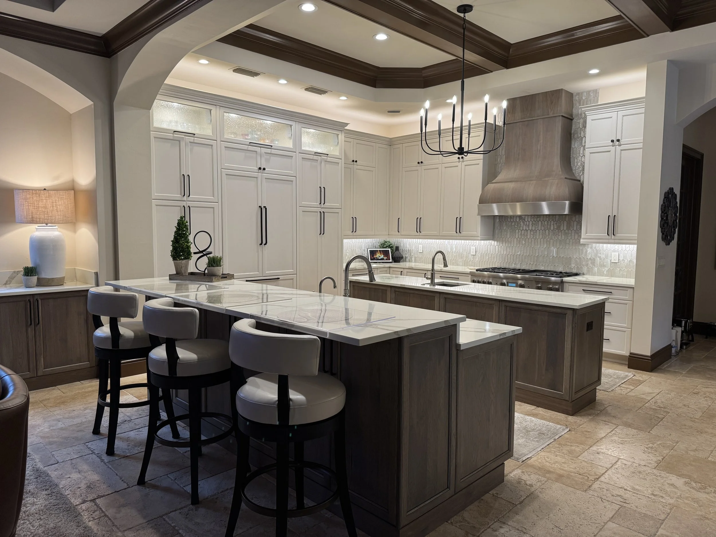 A modern kitchen with white cabinets, a large center island with white marble countertop, stainless steel range hood, black and white stools around the island, a wine rack, decorative plants, a table lamp on a side table, and a chandelier hanging fro