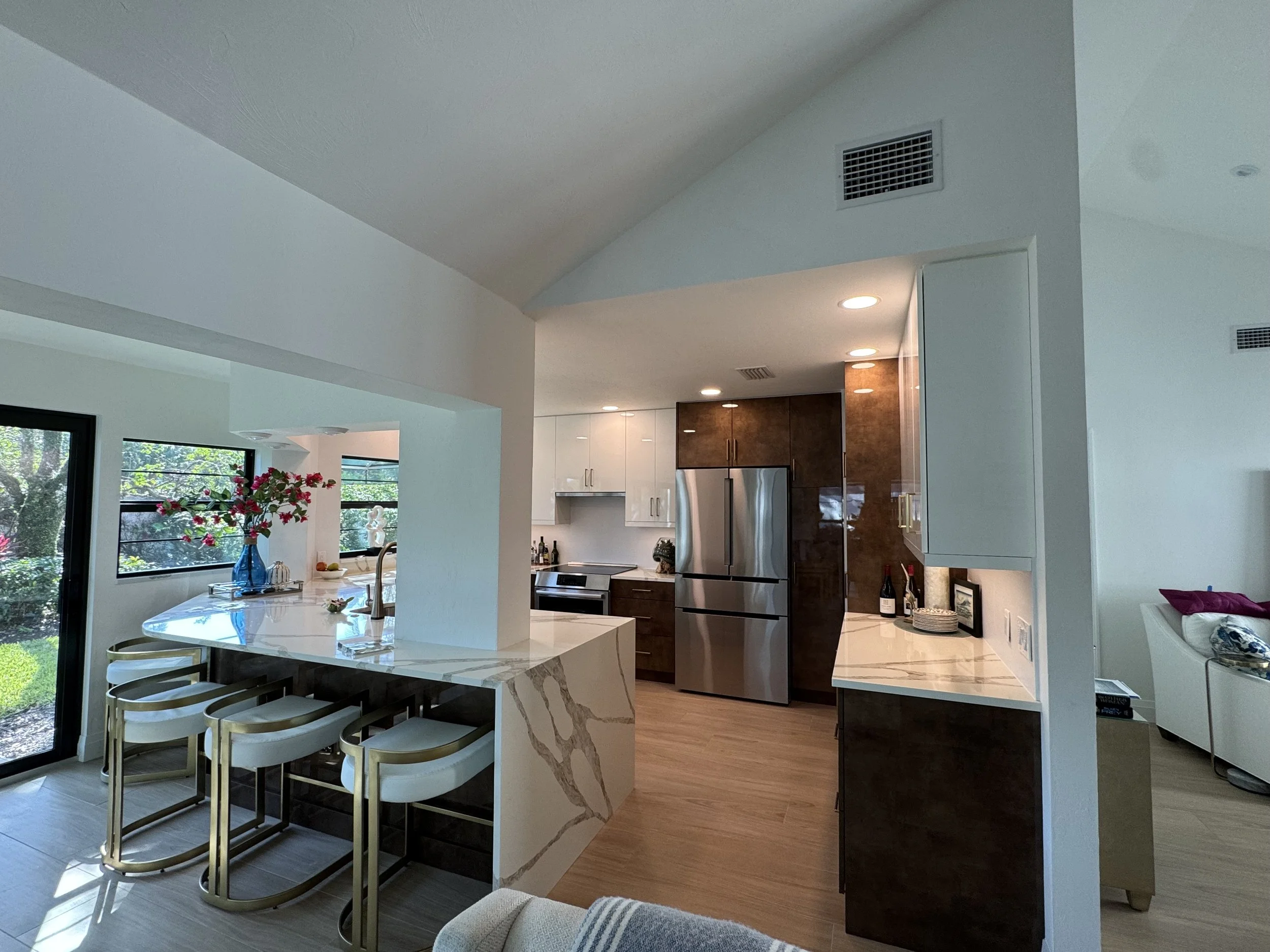 Modern kitchen with white and dark wood cabinetry, stainless steel appliances, a marble island with bar stools, and large windows overlooking greenery.