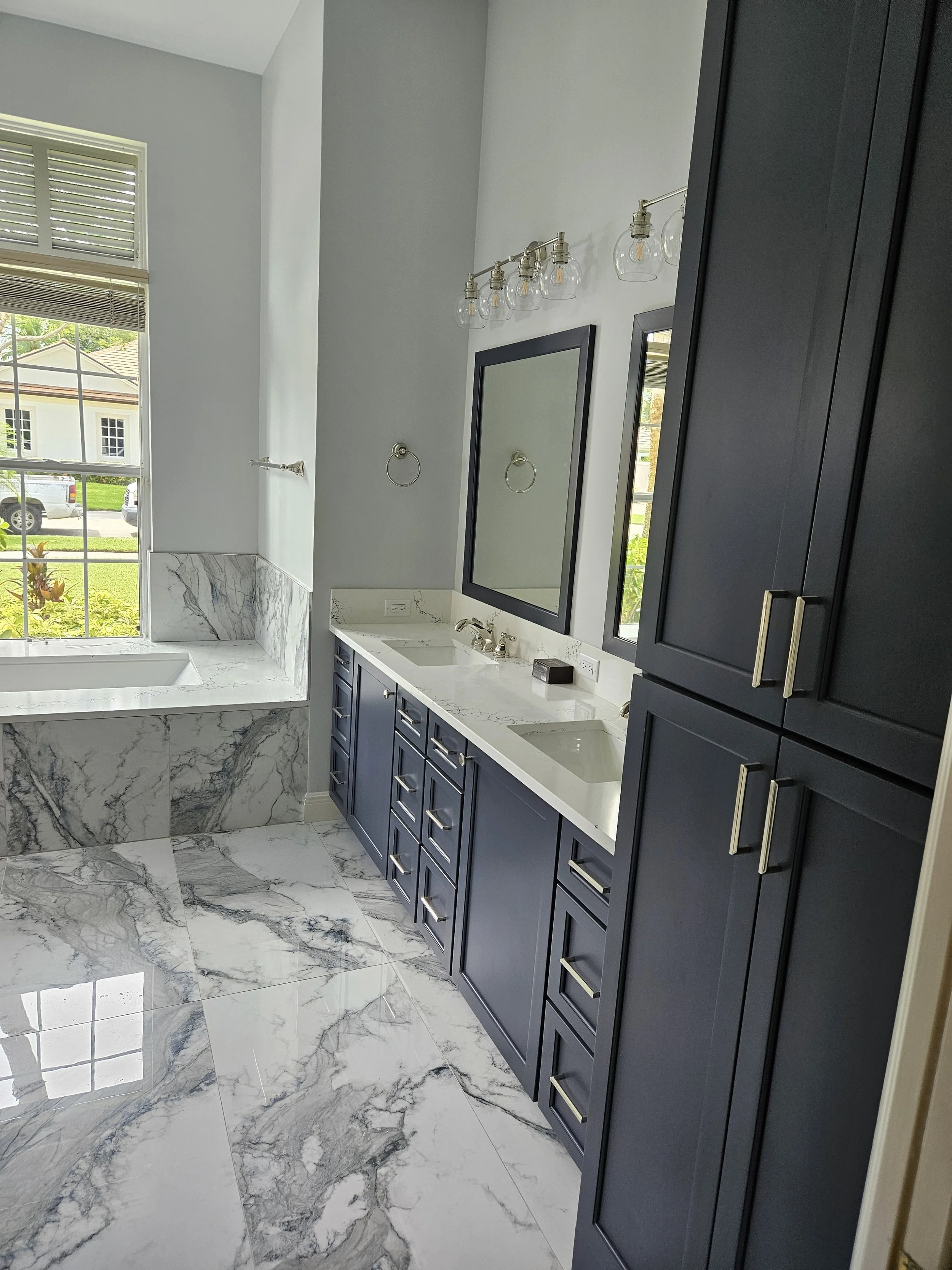 A modern bathroom with a marble bathtub, dark blue cabinets, and elegant fixtures. Large window with blinds, marble floors, and a mirror above the sink.