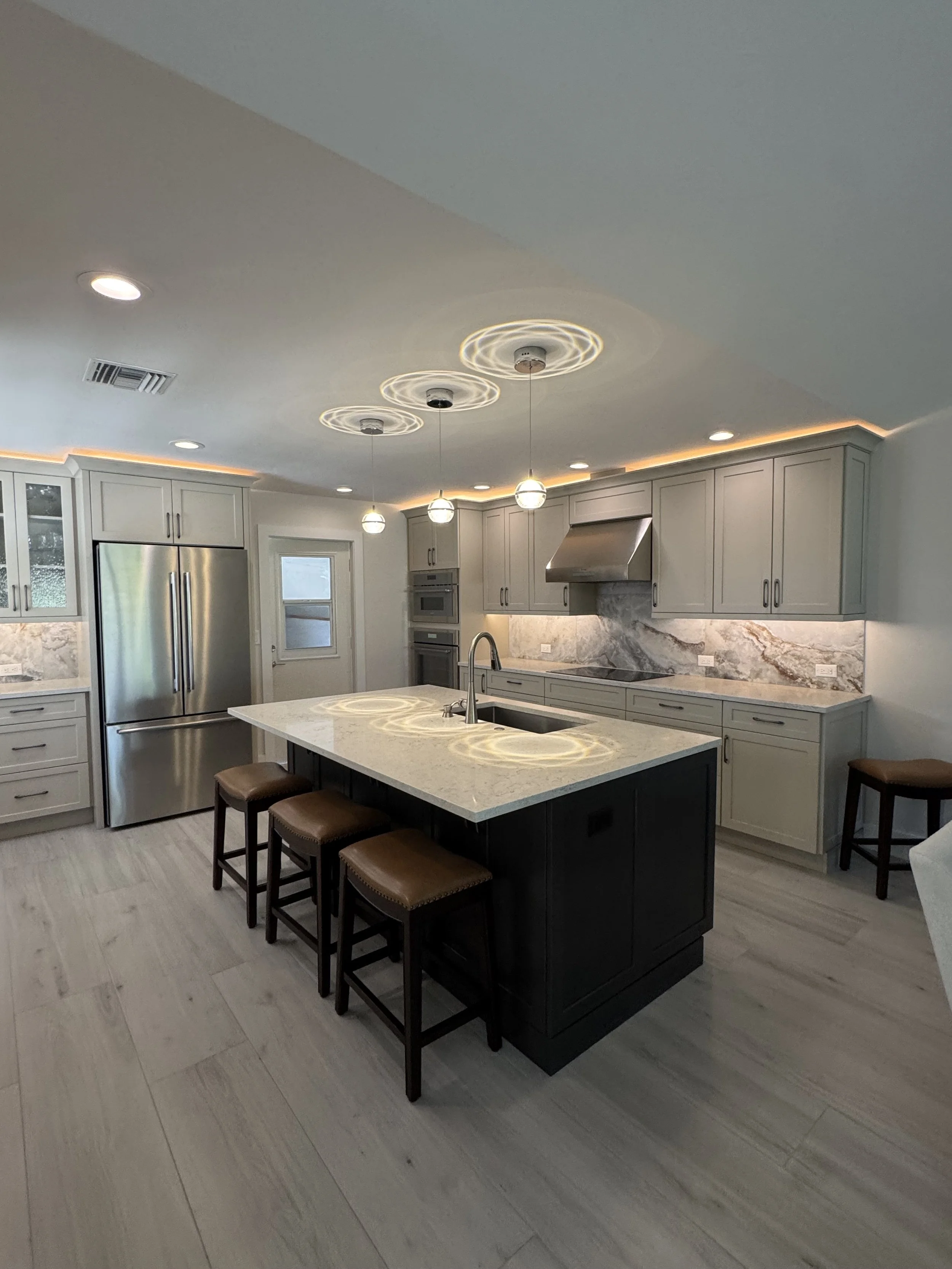 Modern kitchen with light gray cabinets, stainless steel refrigerator, marble backsplash, island with a black base, and three pendant lights hanging above. Four stools are placed around the island, and there's a window and a door on the wall.