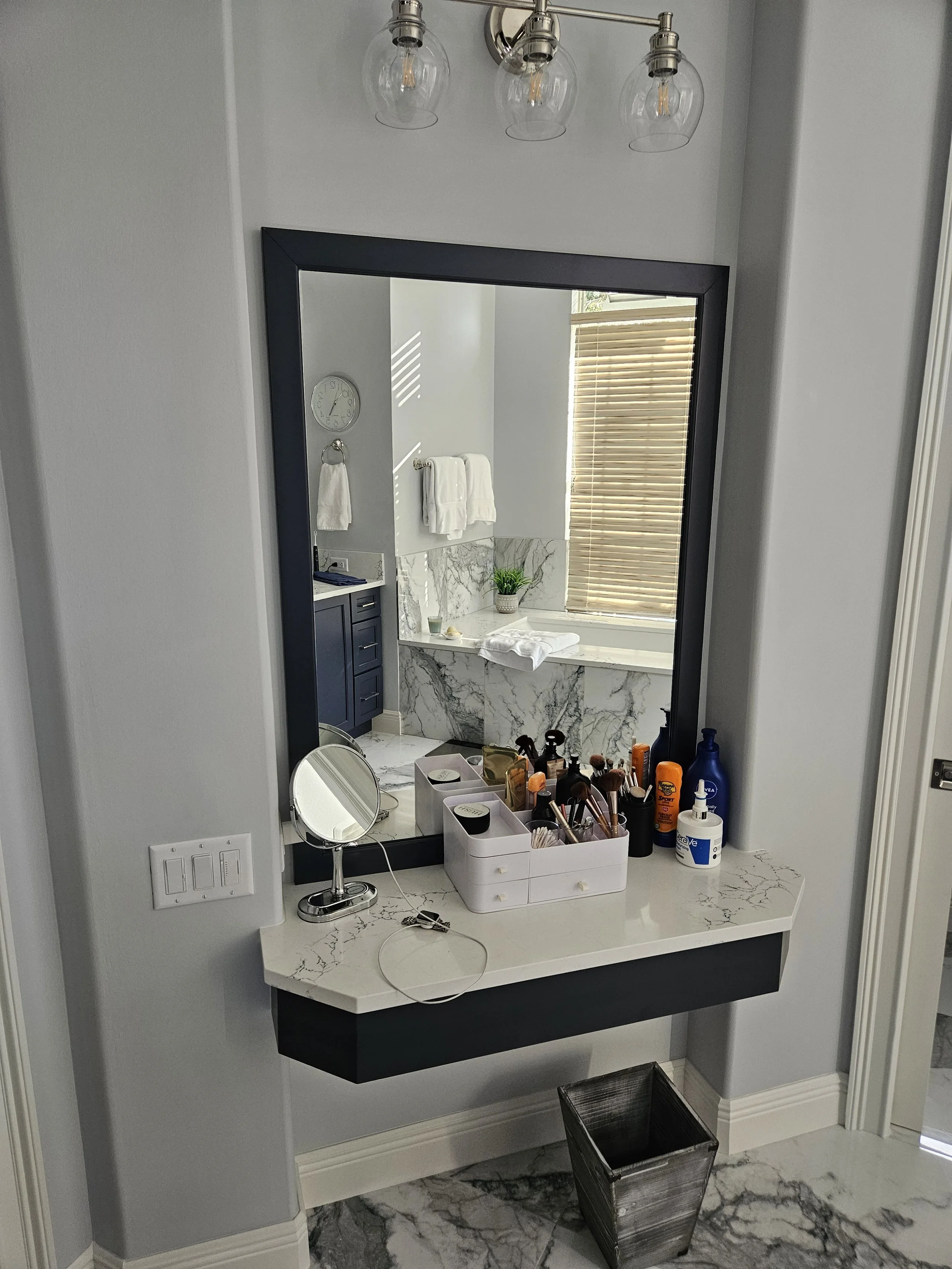 A bathroom vanity mirror with three small containers of makeup brushes, various bottles of lotion, and a small magnifying mirror. The bathroom has a marble countertop with a plant and a towel, two towels hanging on the wall, a window with wooden blin
