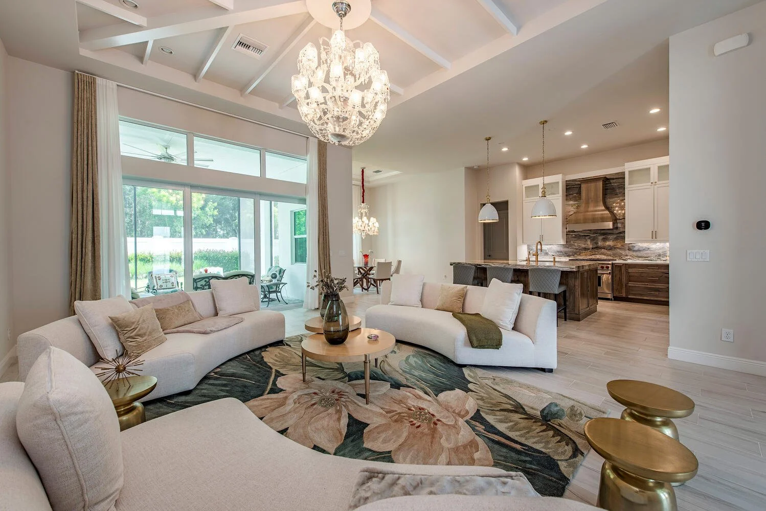 Open-concept living room and kitchen with modern decor, white sofas, floral area rug, chandelier, pendant lights, wooden kitchen island, and large windows showing outdoor greenery.