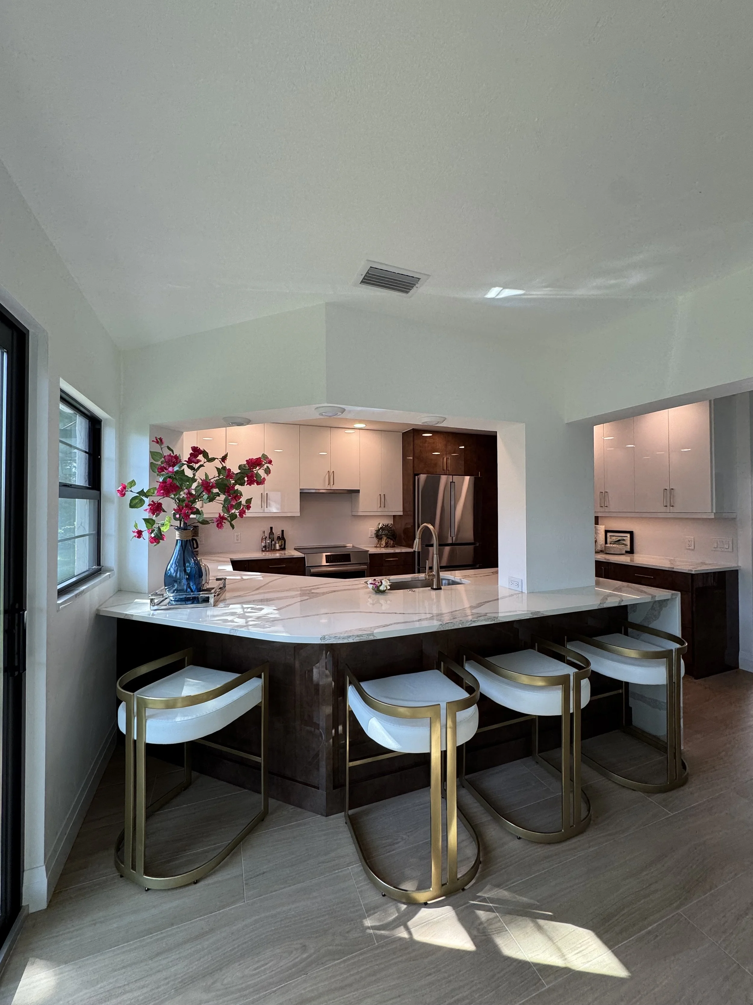 Modern kitchen with a marble island, four bar stools with gold frames, a blue vase with pink flowers, upper white cabinets, and stainless steel appliances under a ceiling with air vents and recessed lighting.