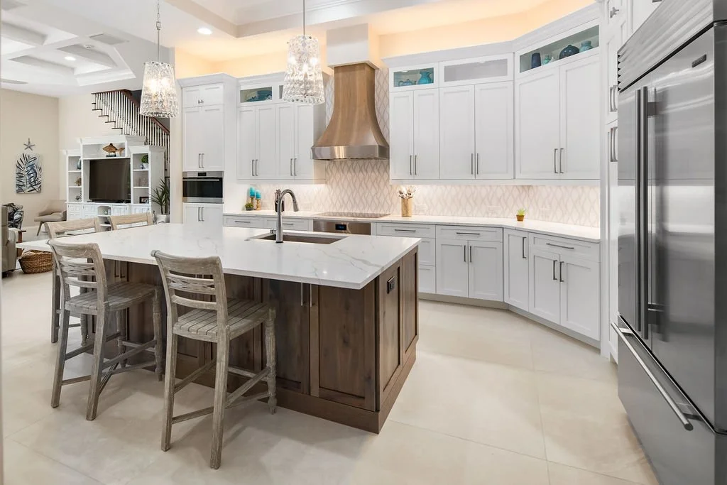 Modern kitchen with white cabinets, a large central island with a marble countertop and wooden base, three rustic wooden chairs, a stainless steel range hood, and built-in appliances, with a cozy living area visible in the background.